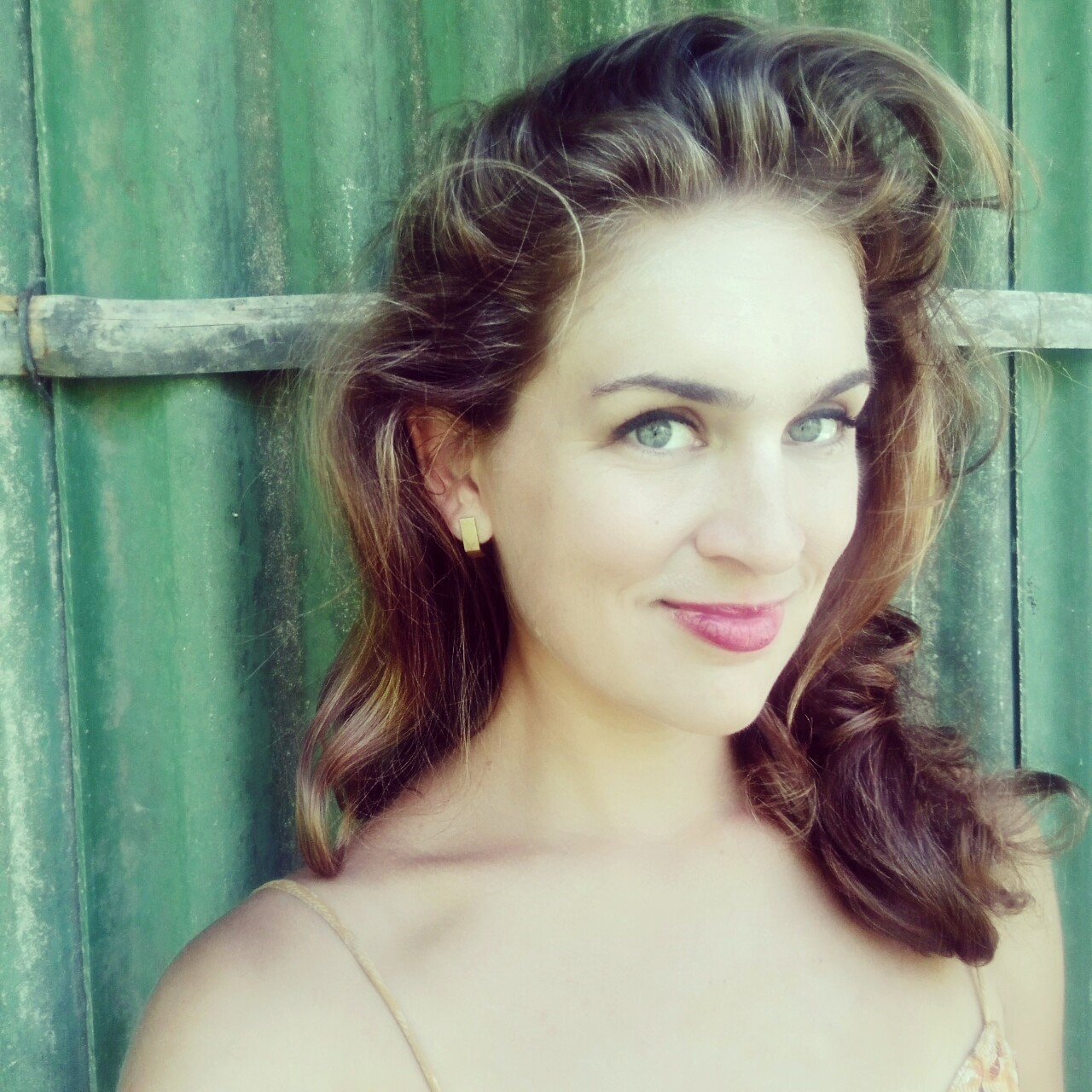 A woman with curly brown hair and blue eyes smiling, standing against a weathered green wooden background.