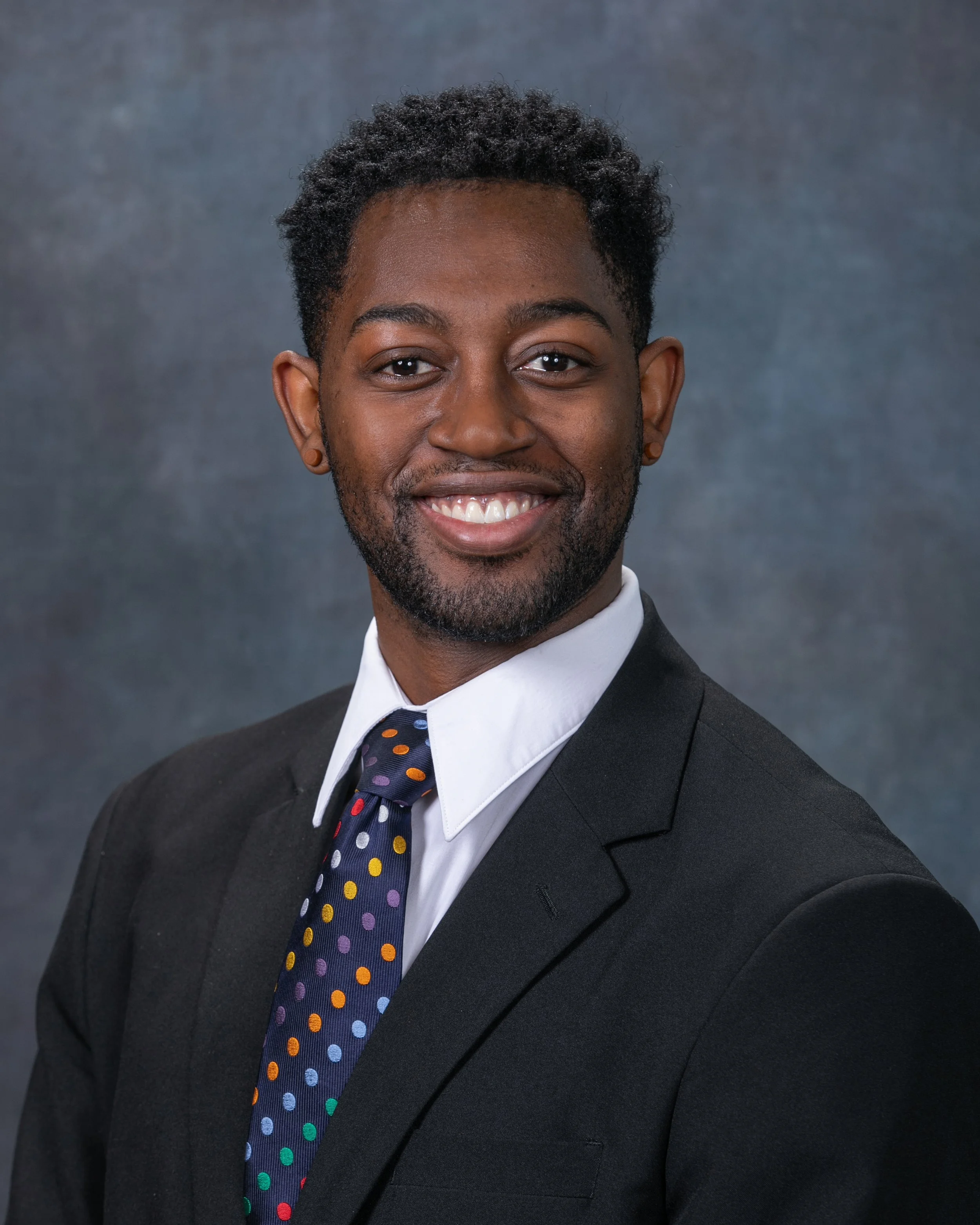 A young man smiling, wearing a dark suit, white shirt, and a navy tie with multicolored polka dots, against a gray textured backdrop.