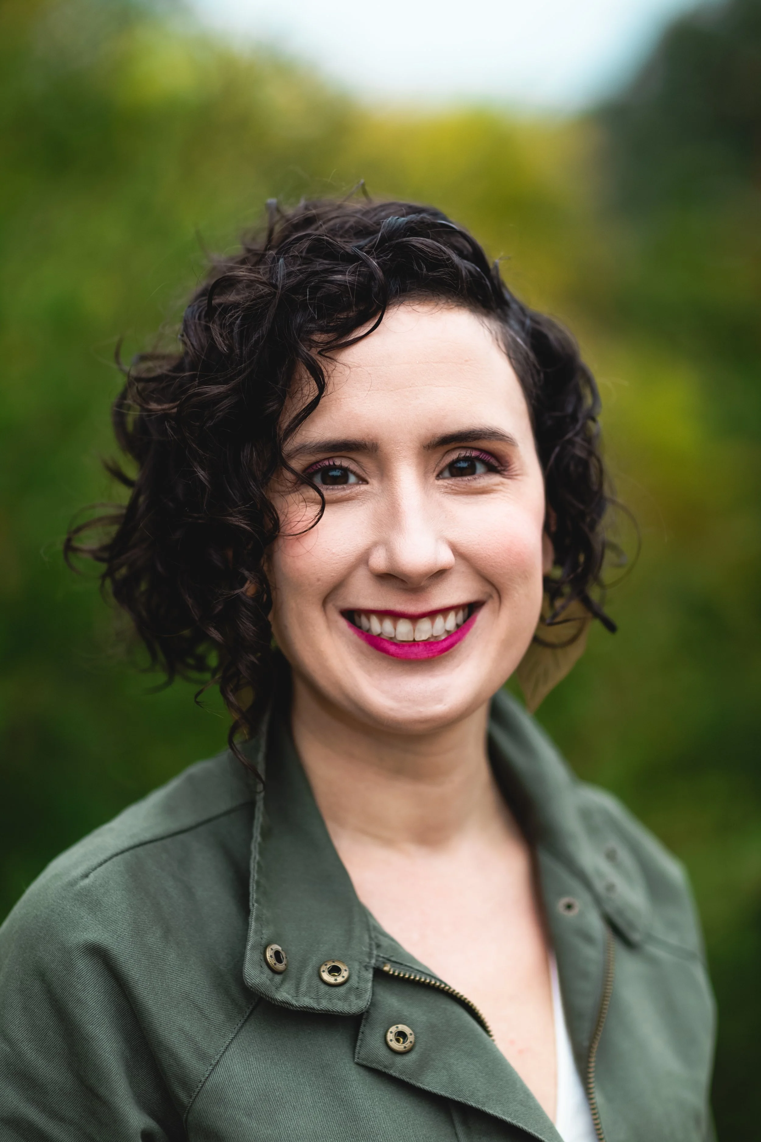Close-up of a woman with curly dark hair, smiling, wearing a green jacket outdoors with blurred green trees in the background.