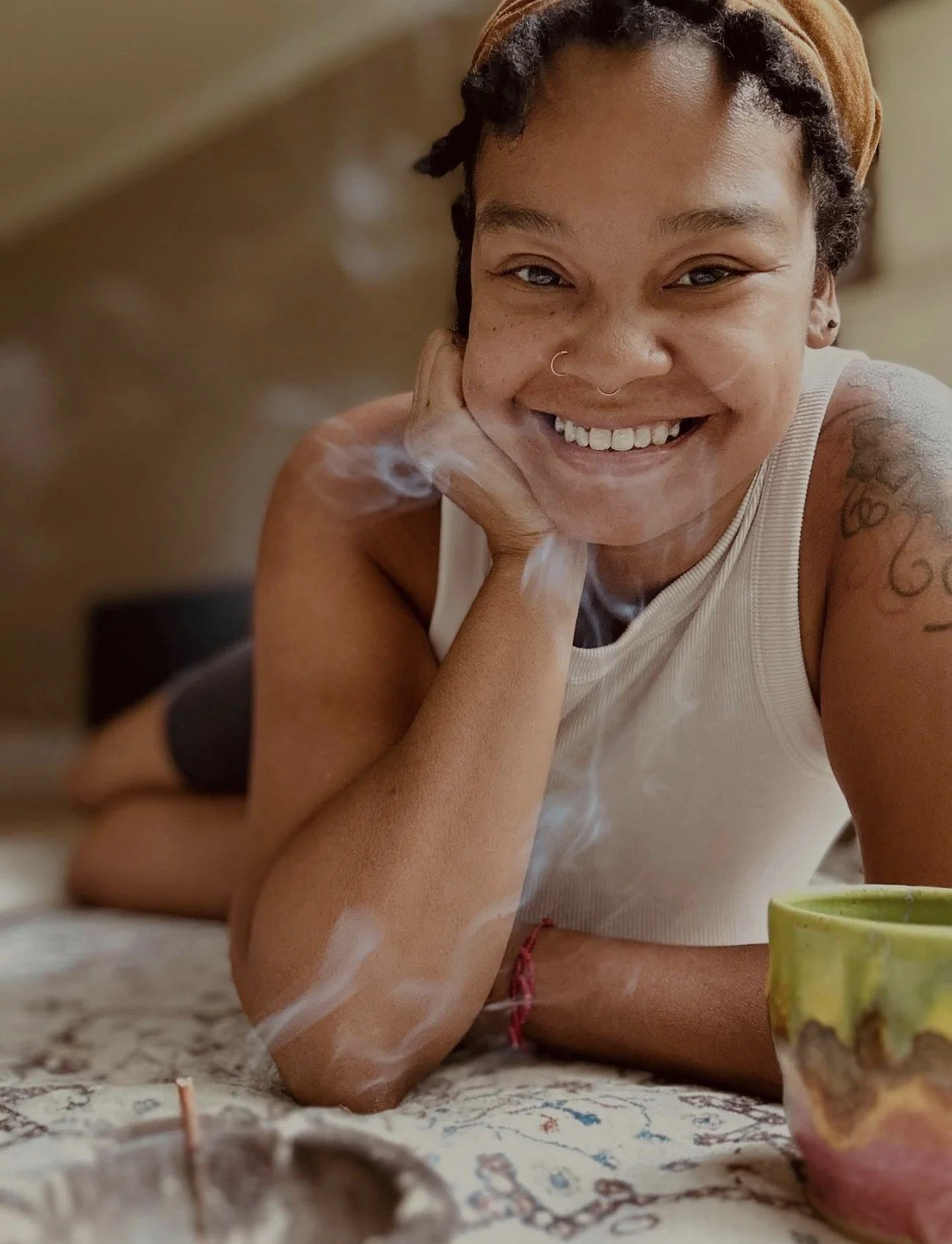 Young woman with short curly hair, tattoos on arm, nose ring, smiling and lying on her stomach on a carpet with a colorful mug nearby.