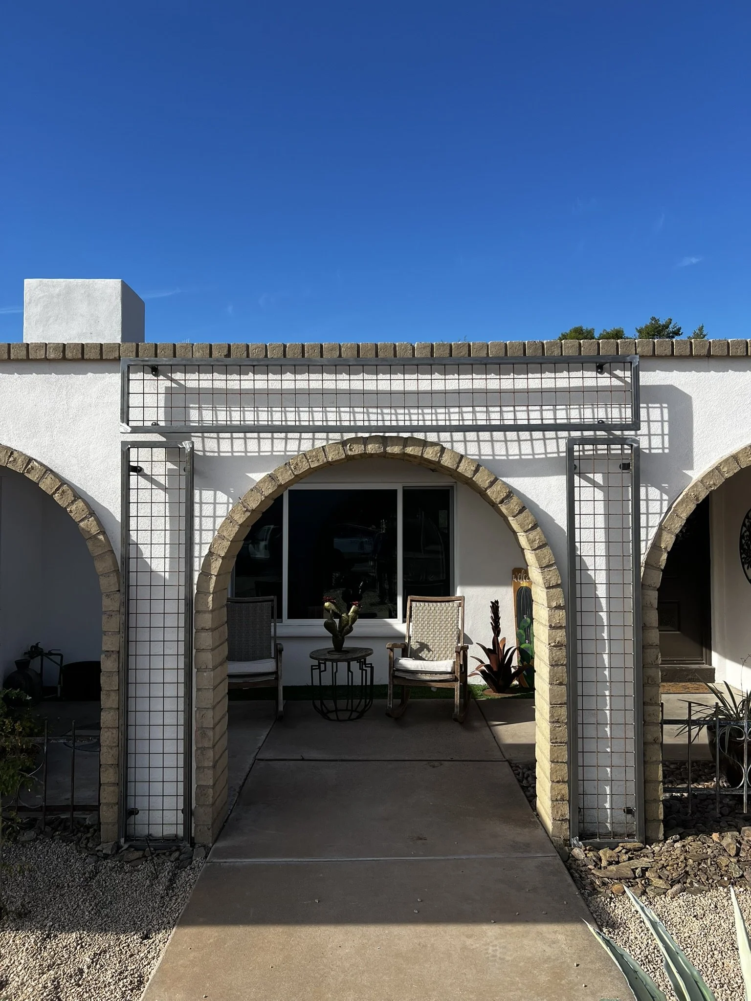 Front view of a white building with arched doorways, decorative metal latticework, outdoor chairs, potted plants, and a sidewalk under a clear blue sky.