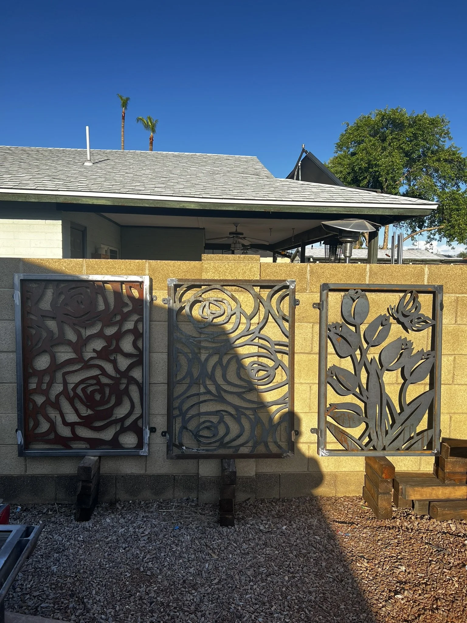 Three decorative metal panels with floral designs in front of a yellow brick wall, with a house, trees, and clear blue sky in the background.