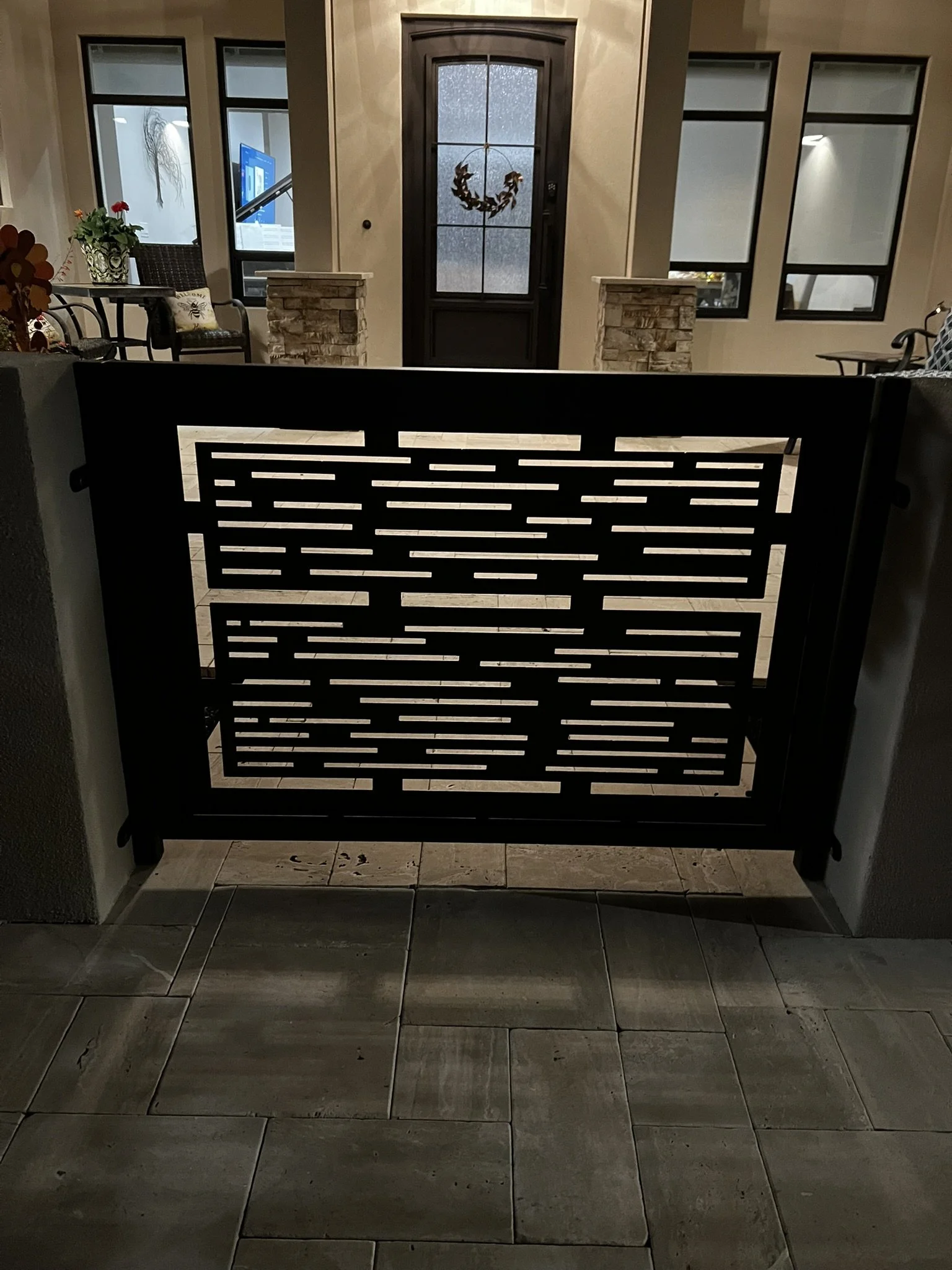 An outdoor front porch area illuminated at night, featuring a black metal gate in the foreground, a front door with a decorative wreath, and a windowed wall with stone accents.