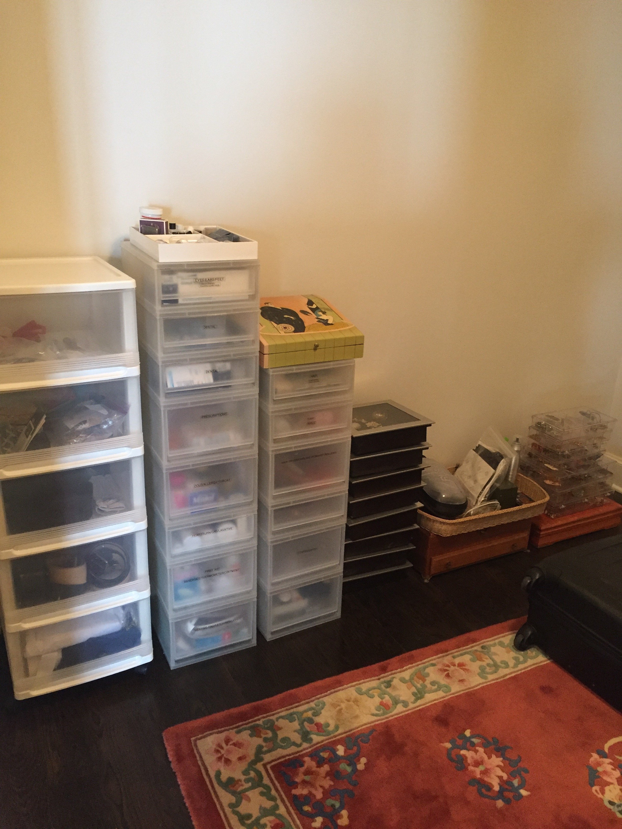 A collection of plastic storage drawers and containers filled with various items, placed against a beige wall in a room with a dark wood floor and a colorful area rug.