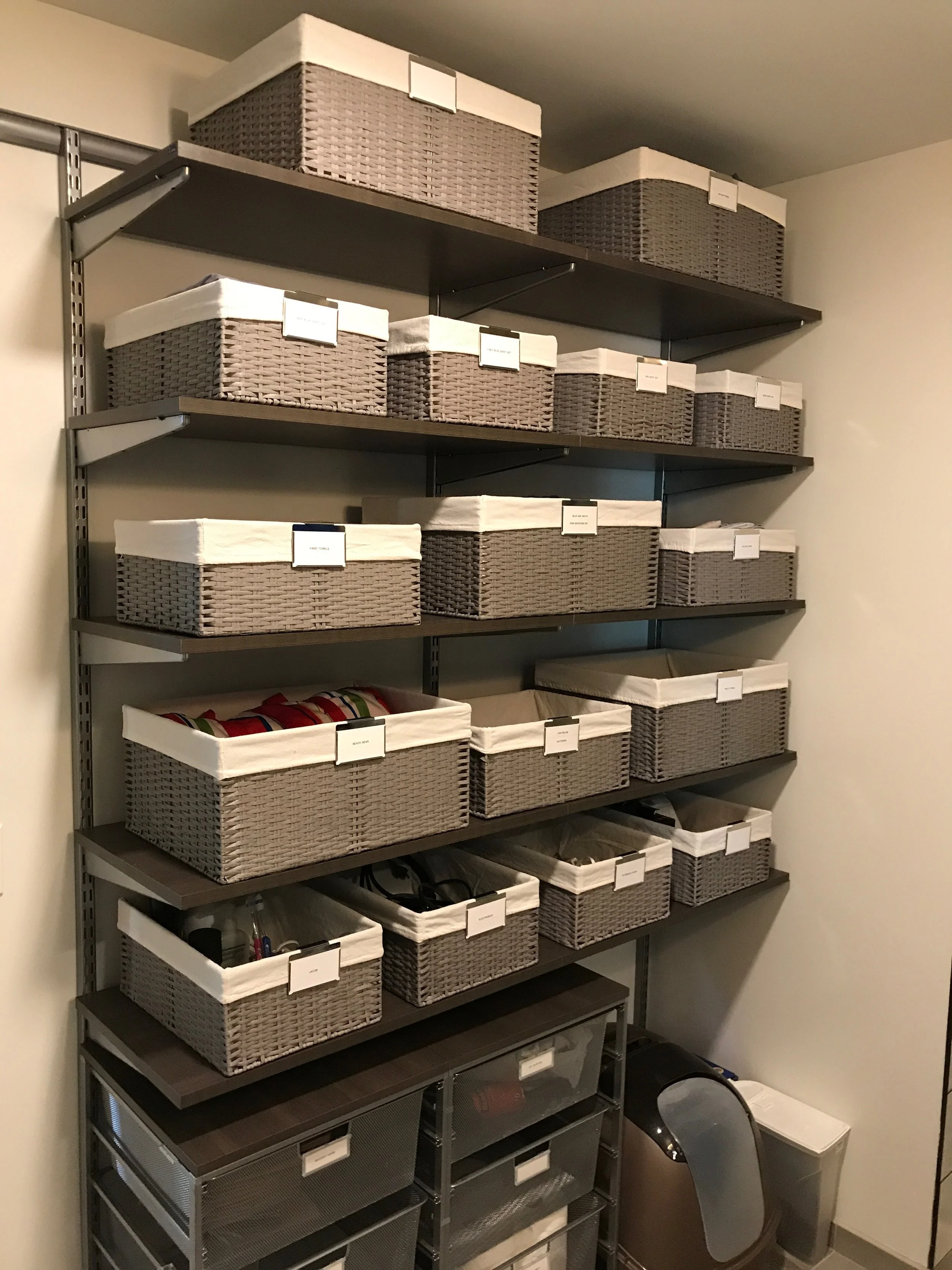 Shelves filled with various woven baskets and storage bins, some labeled, in a closet or storage room.
