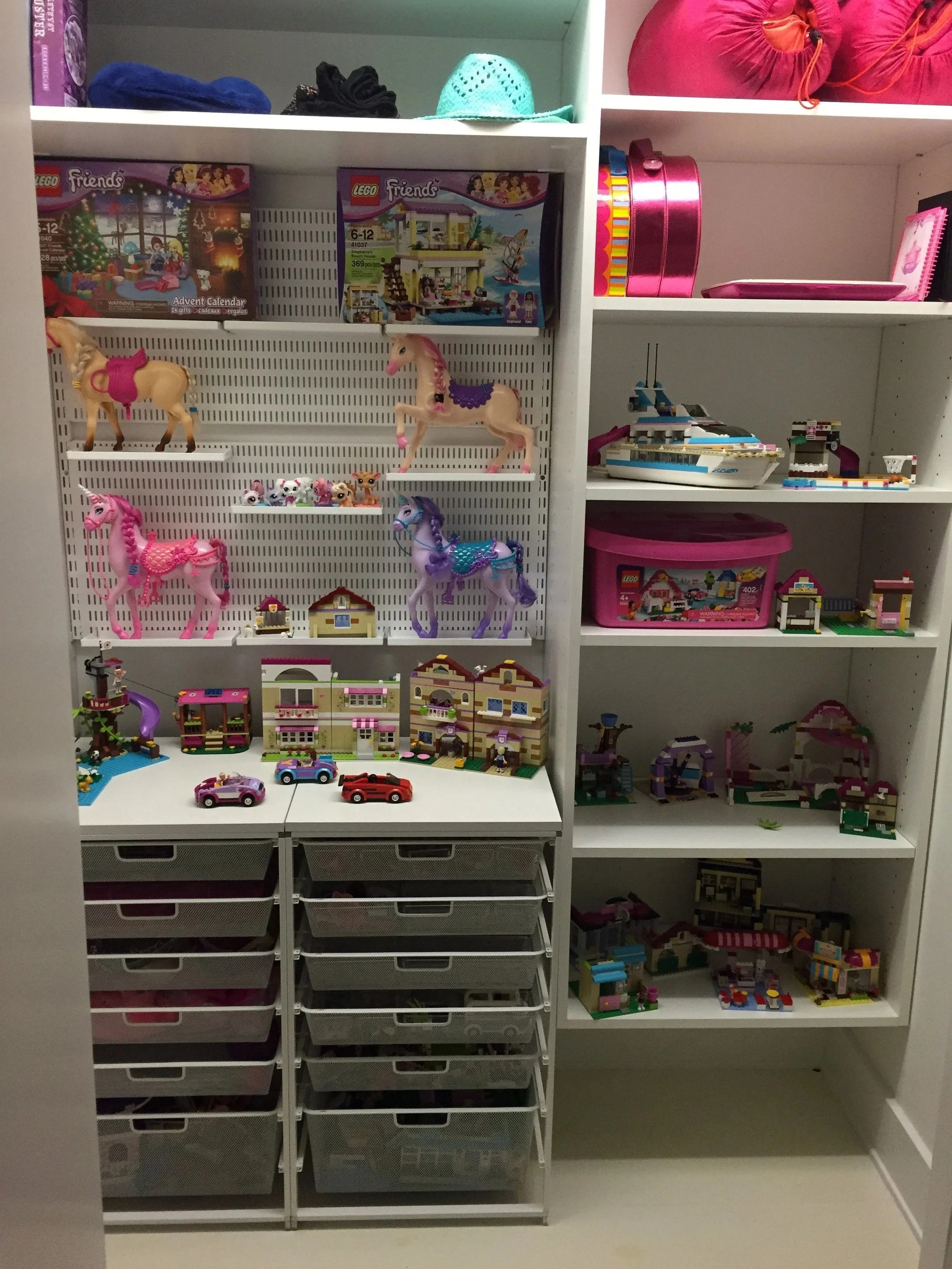 A white storage unit filled with toys, including Lego sets, horse figurines, and miniature vehicles, with plastic drawers at the bottom and various other toys on the shelves.