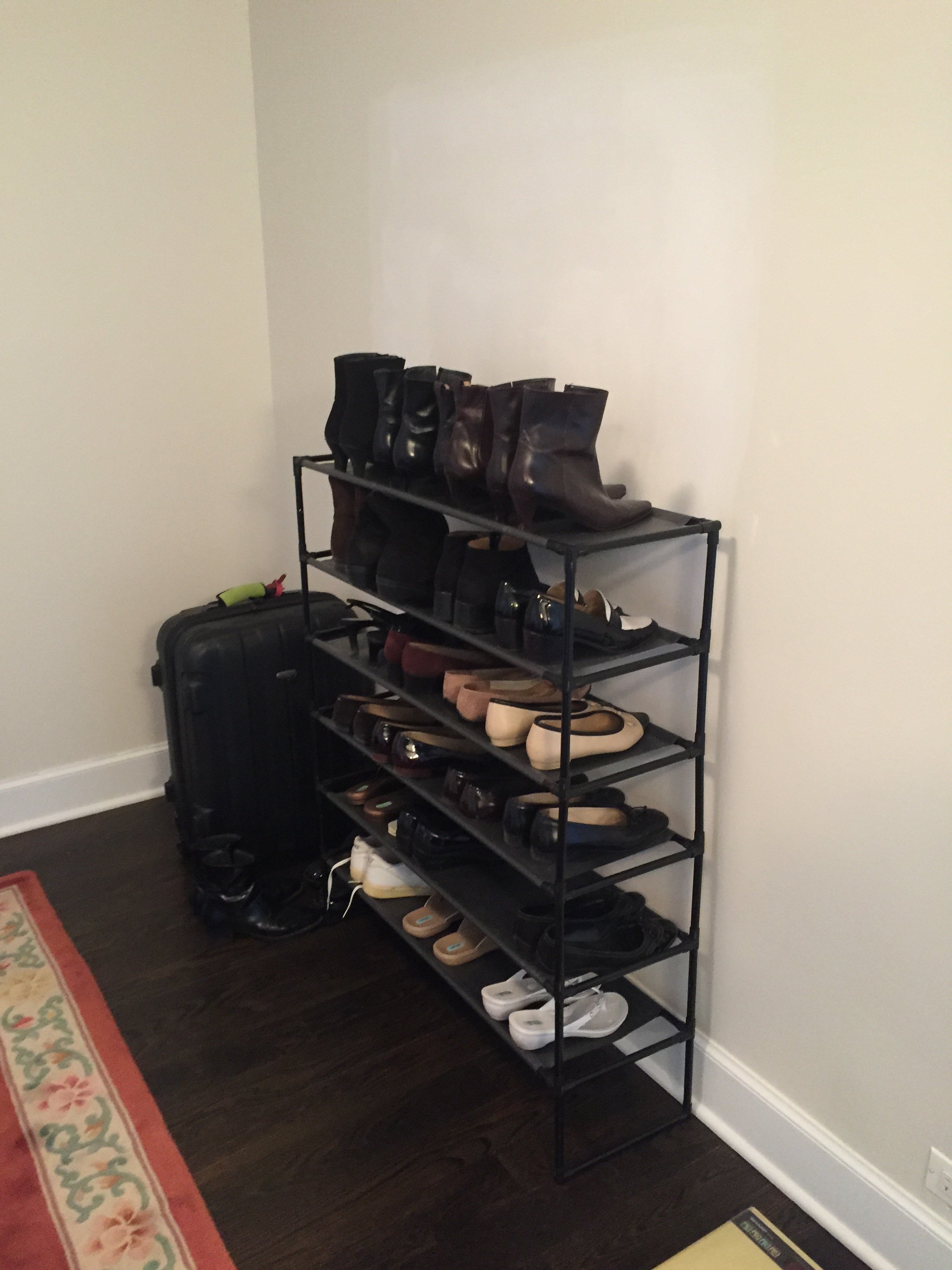 A black metal shoe rack with six shelves, filled with various pairs of shoes including boots, flats, sandals, and sneakers, positioned against a plain white wall in a room with dark wooden flooring.