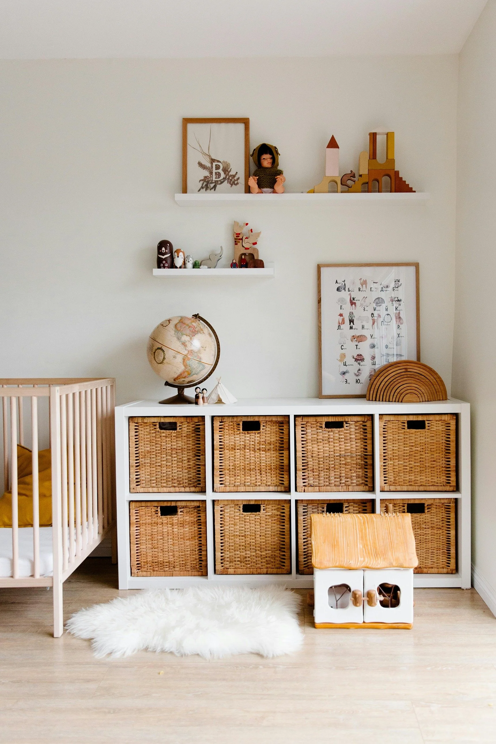 Organized children's playroom or nursery with a white storage unit with woven basket drawers, decorative items on top including a globe and small toys, a white rug, a dollhouse, and art and collectibles on shelves and wall.