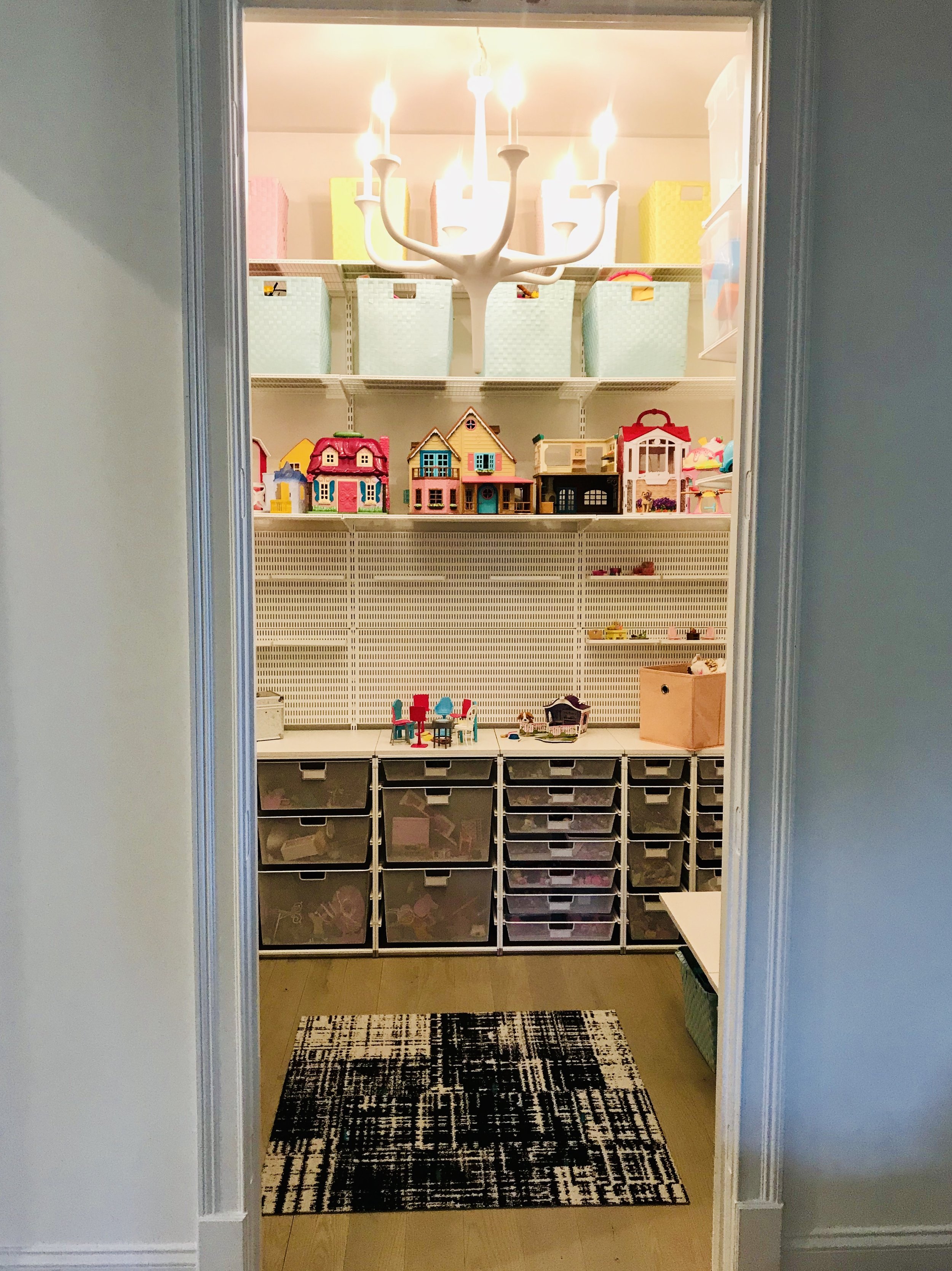 View through doorway into a playroom with a white chandelier, shelves full of toy houses, and storage bins for toys with a black and white rug on the floor.