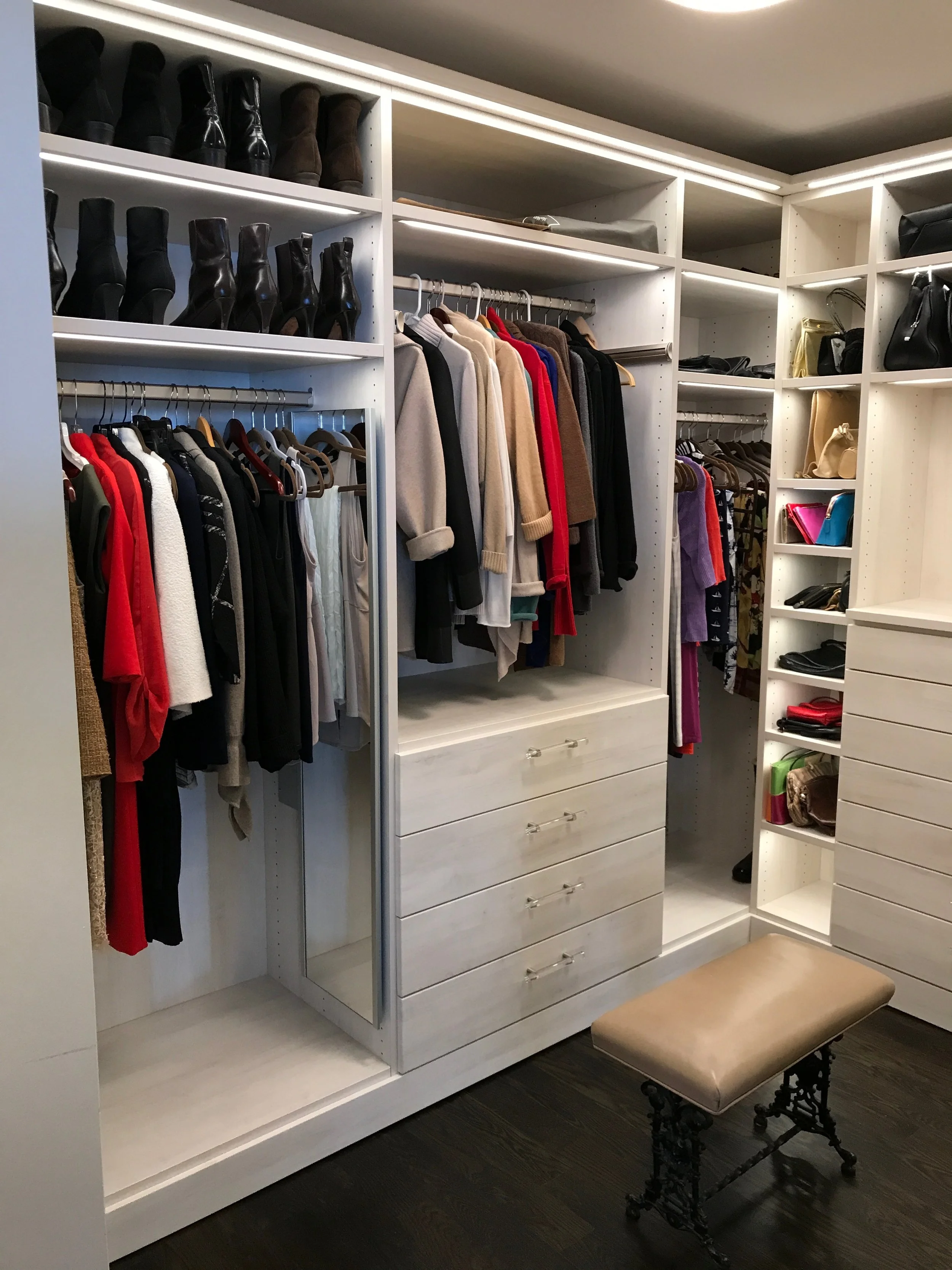 Walk-in closet with white shelving containing shoes, handbags, and clothing. Clothes on hangers include sweaters and jackets. There is a beige ottoman with black ornate legs on a dark hardwood floor.