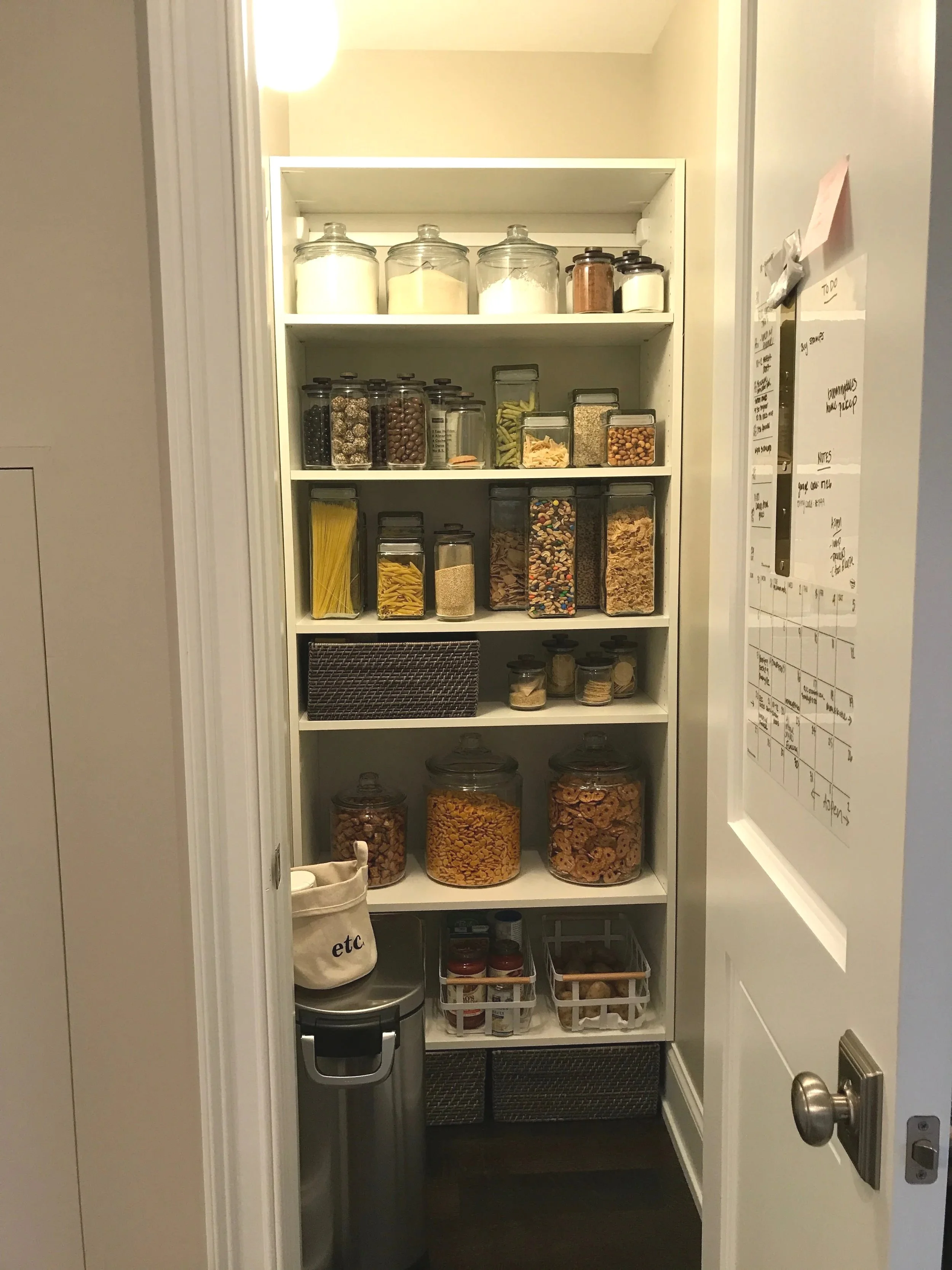 A pantry with white shelves filled with glass jars containing pasta, cereal, beans, spices, and snacks.