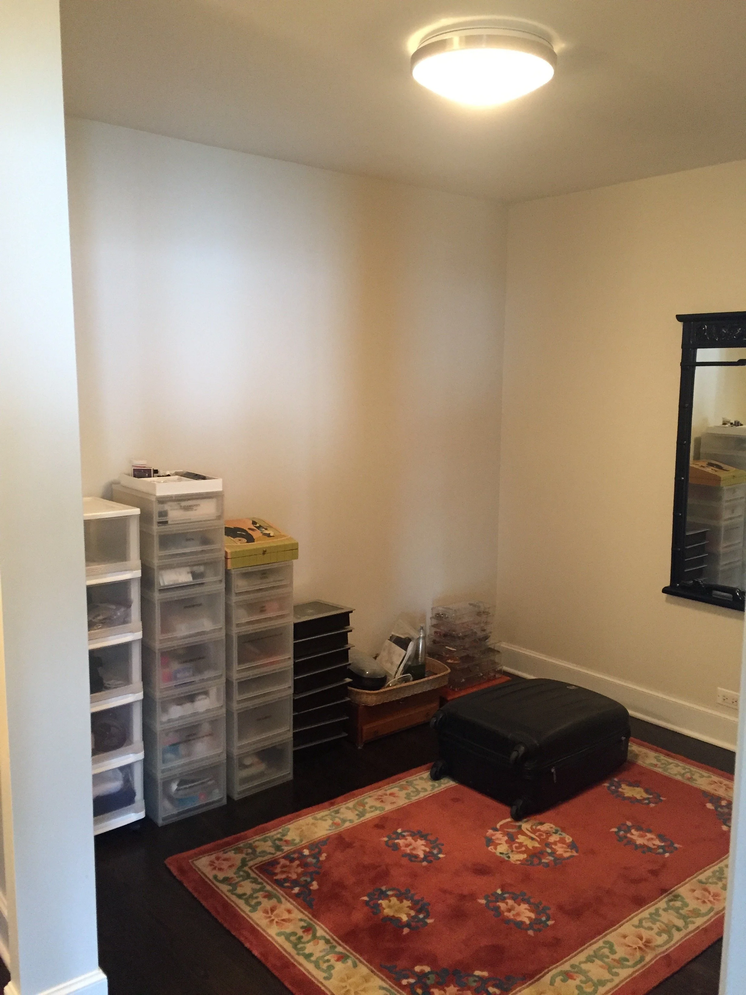 A small room with a rectangular red and beige floral area rug, plastic storage drawers, a black suitcase, and a mirror on the wall.
