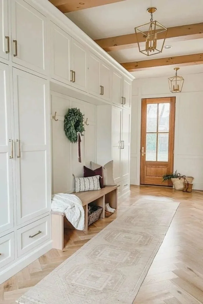 Entryway with white built-in cabinets, a wooden bench with pillows, a green wreath hanging on the wall, a long beige rug, a glass-pane door, and two gold geometric pendant lights.