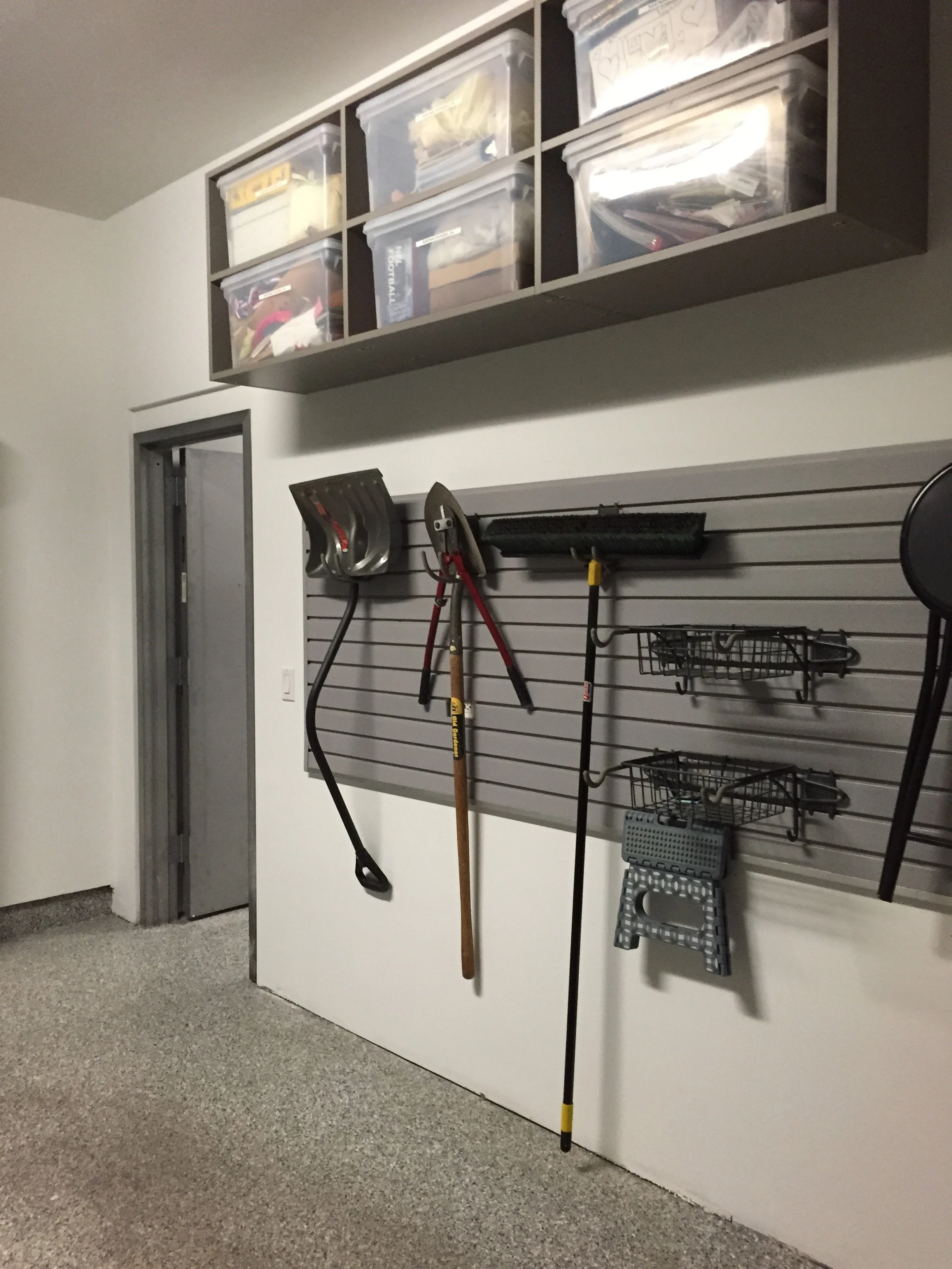 Garage storage area with a slatwall holding garden tools, a step stool, and a shelf with plastic storage bins containing various items.