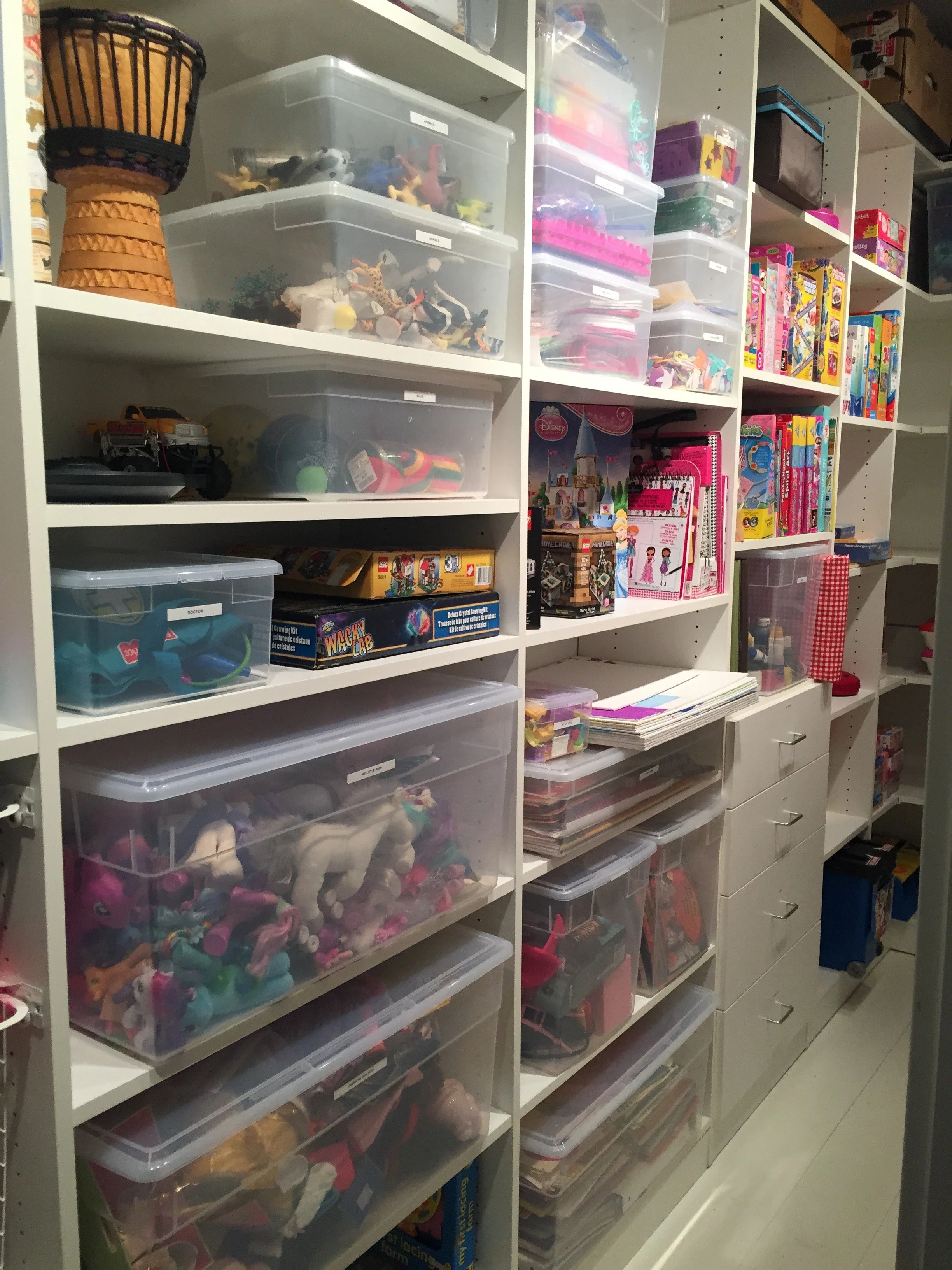 Section of a closet shelves filled with toys, including toy animals, building blocks, and board games in plastic storage containers and on shelves.