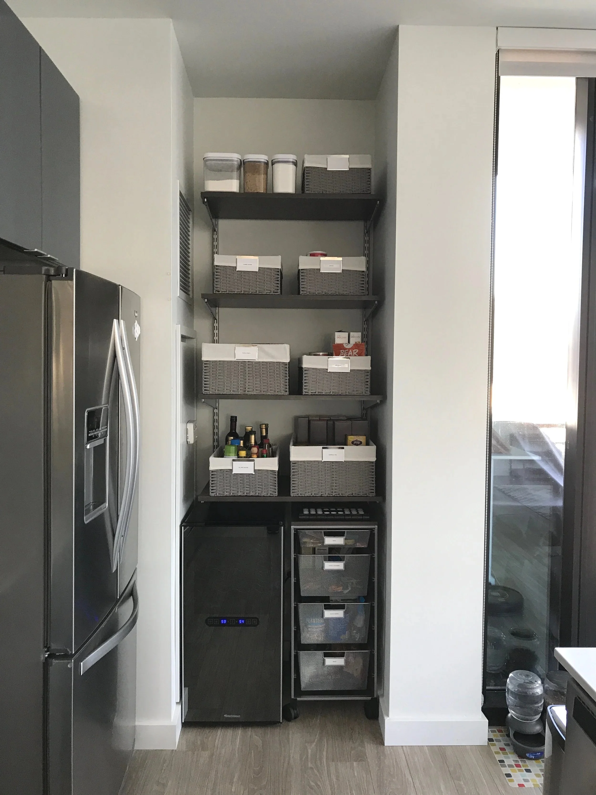 Kitchen storage area with open shelves containing containers, baskets, and bottles, next to a stainless steel refrigerator and a glass sliding door.
