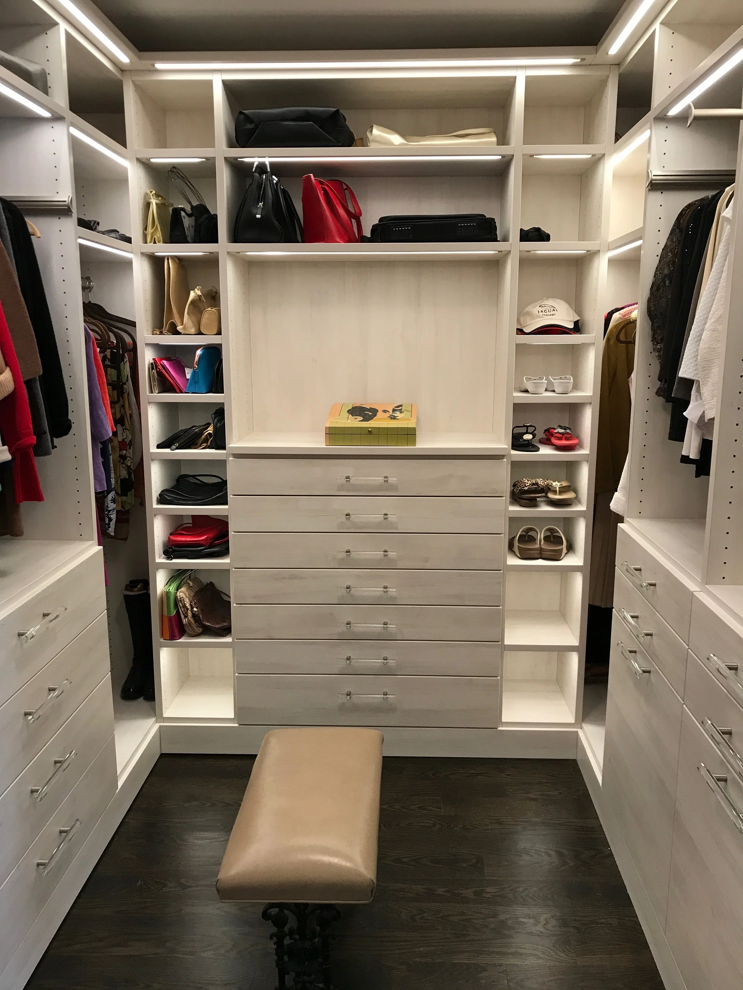 A walk-in closet with white shelving, drawers, and hanging space, containing handbags, shoes, hats, and clothes, with a beige padded stool in the center on dark wooden flooring.