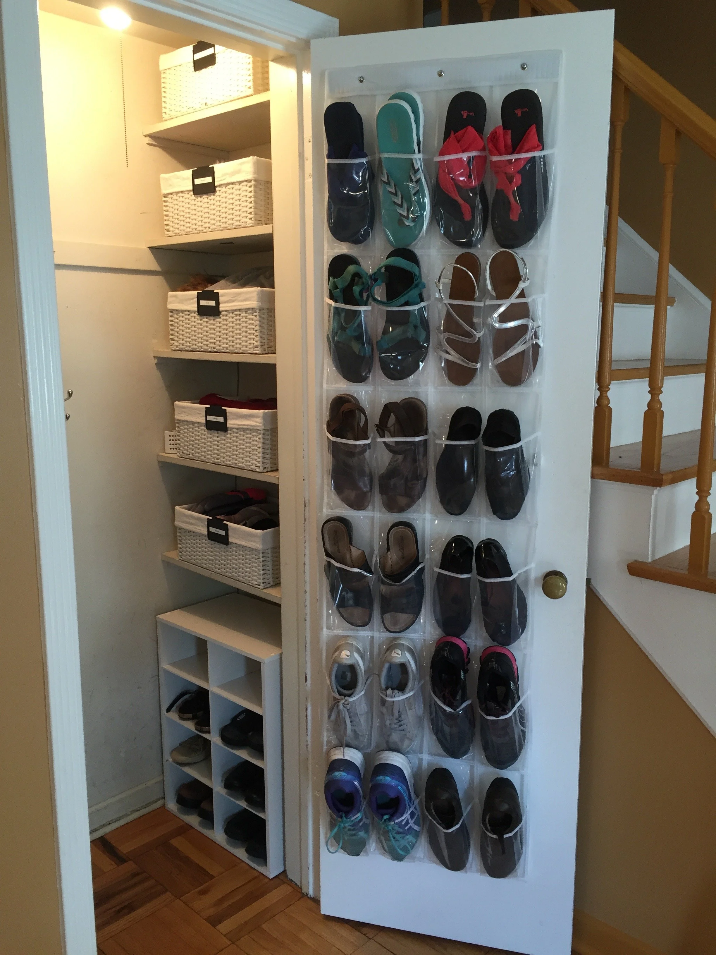 A white door with plastic pockets holding various styles of shoes, including sneakers, sandals, and slippers, next to a small cabinet with multiple pairs of shoes and a closet with shelves and baskets.