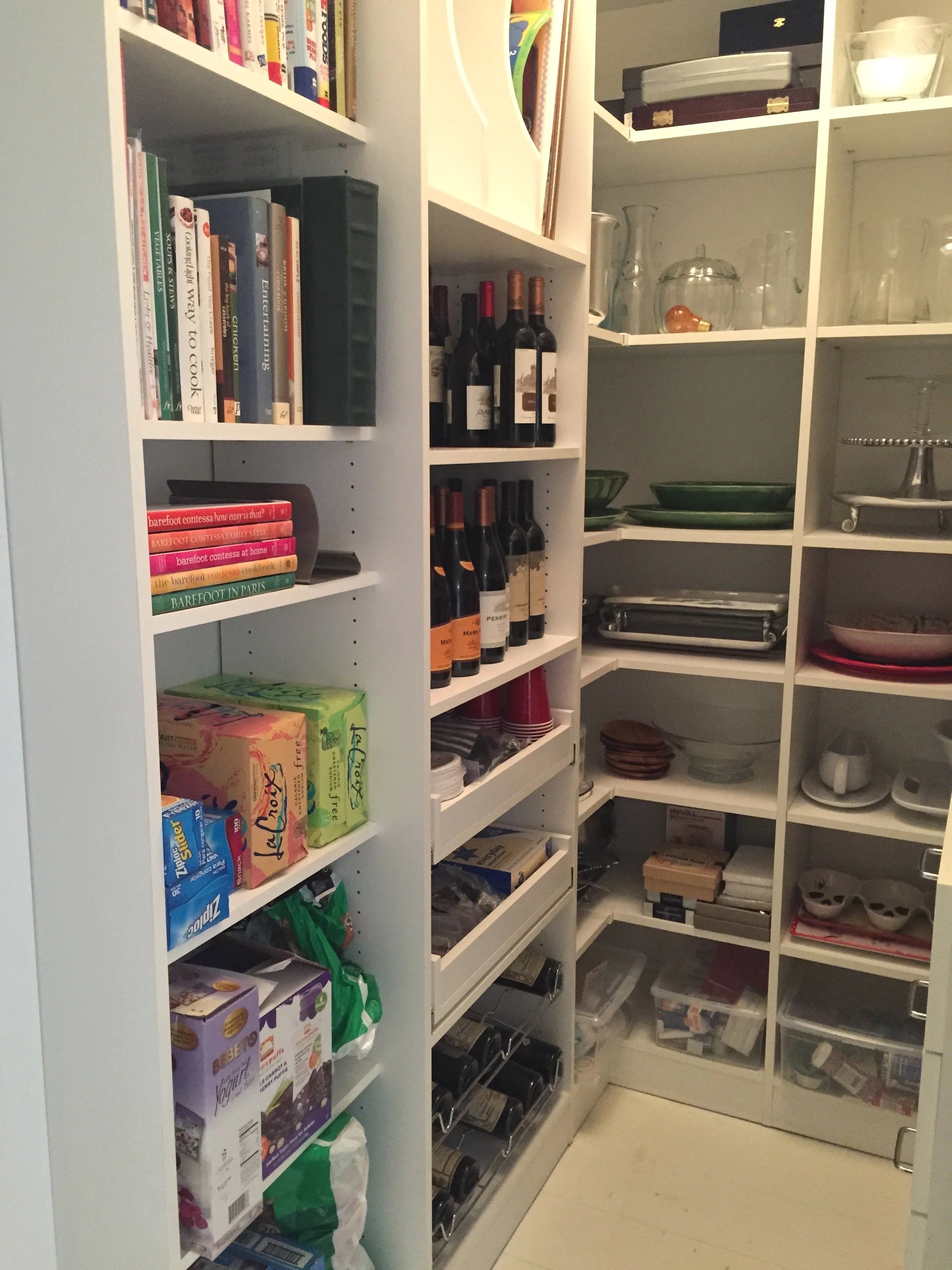 View of a pantry with white shelving filled with food, beverages, dishes, glassware, and storage containers.