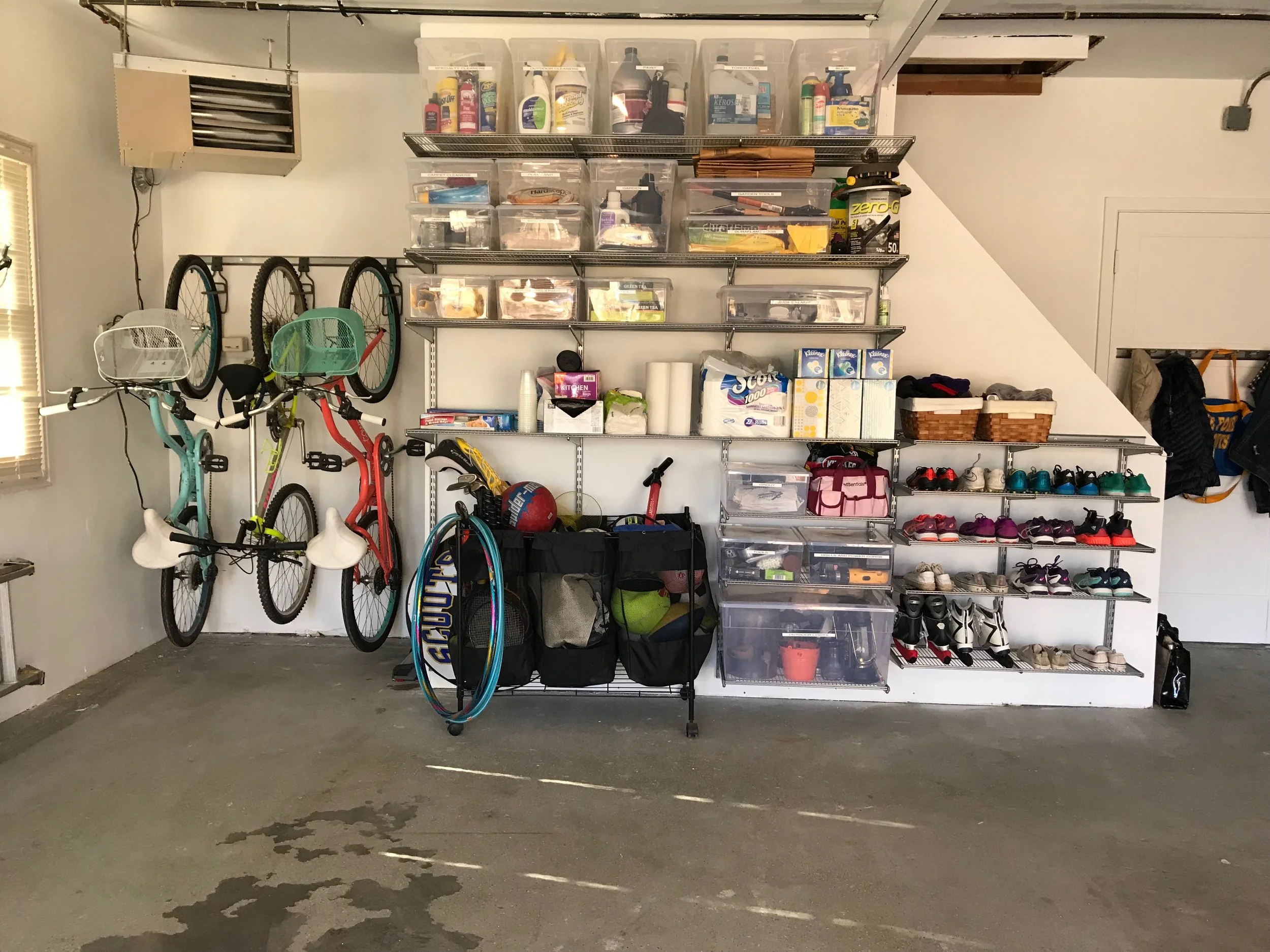 Garage wall with shelves holding storage bins, shelves with baskets, and a row of shoes, including rollerblades, organized by color. Three bicycles are hanging on the wall, and footballs, a jump rope, and bags are on the floor.