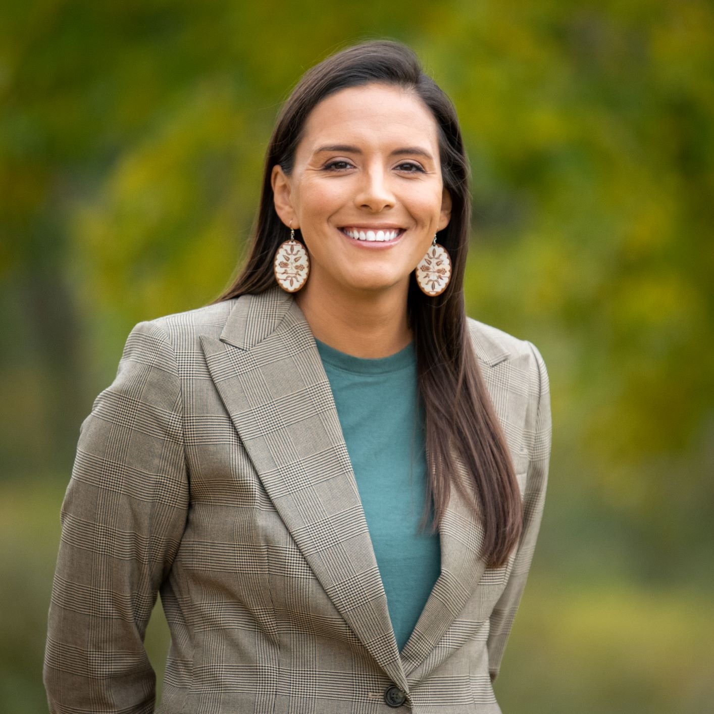A woman with long dark hair smiling outdoors, wearing a plaid blazer, green shirt, and large circular earrings with a tree design, against a blurred background of trees.