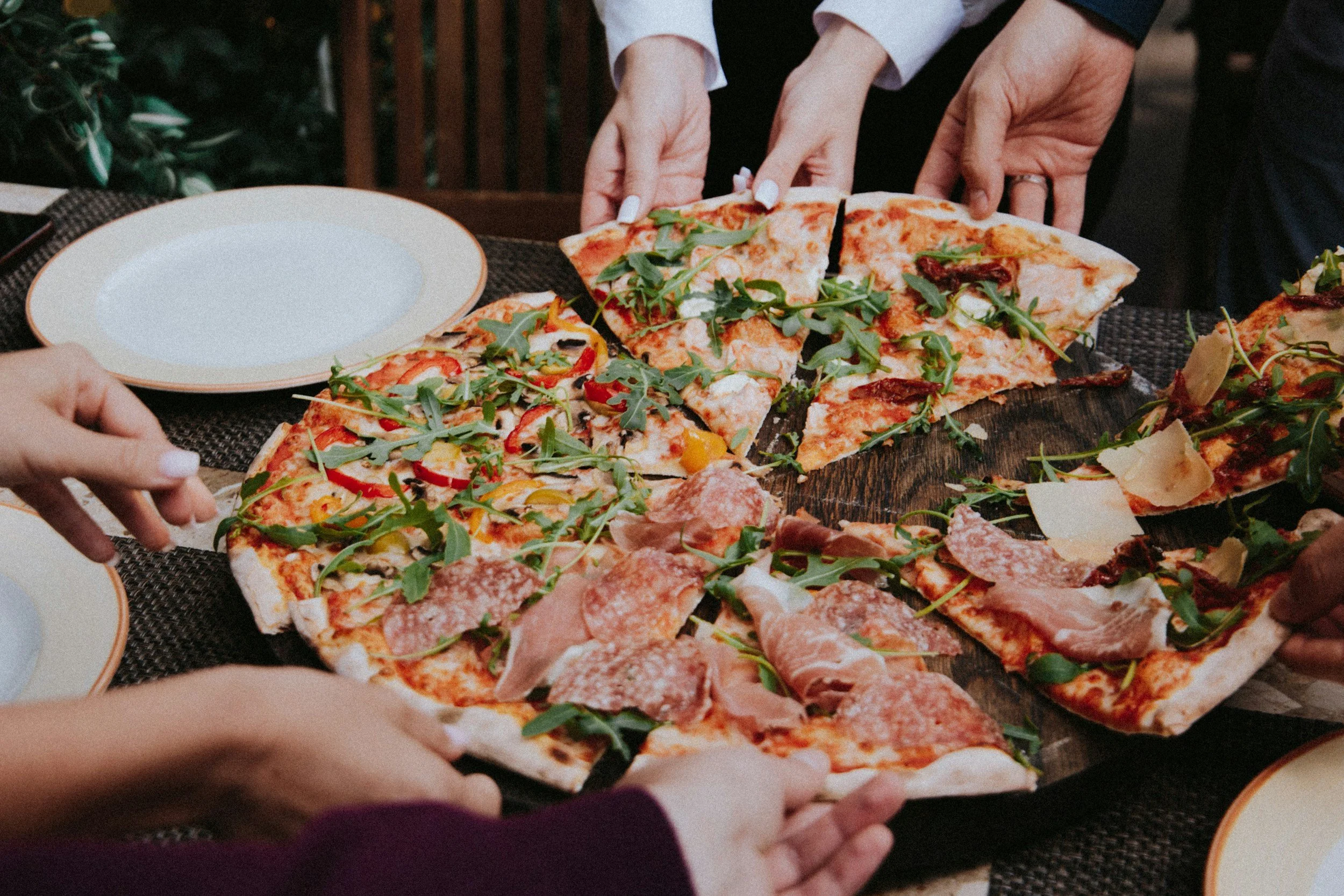 People reaching to serve themselves slices of pizza on a table.
