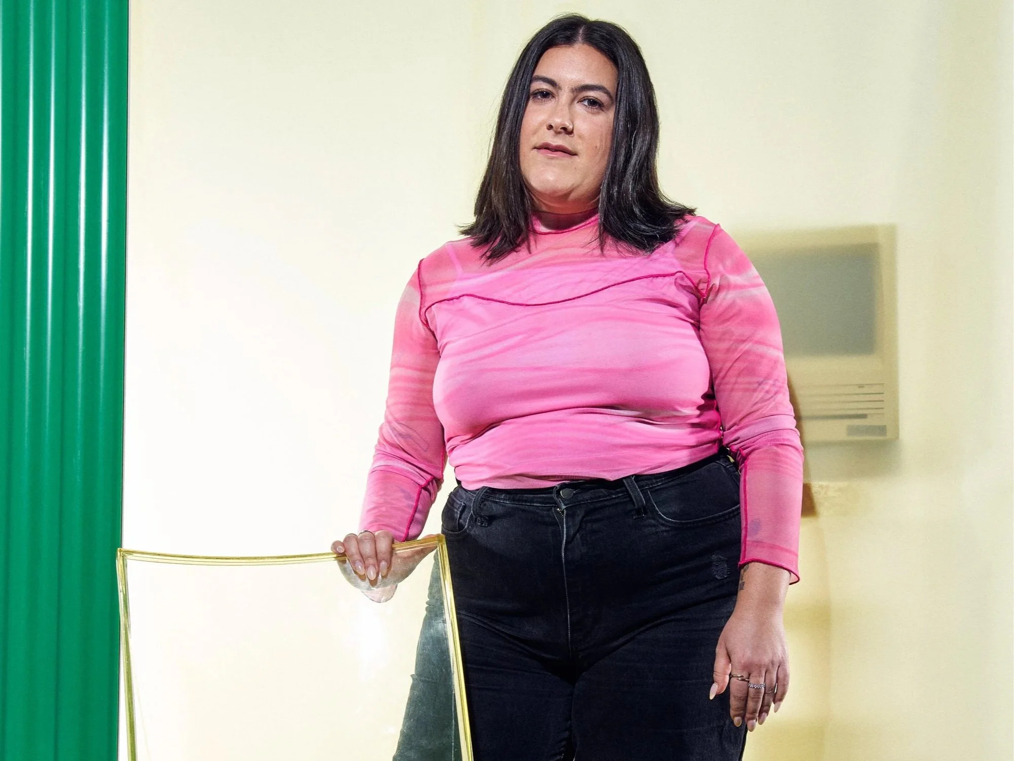 Person with long dark hair wearing a pink shirt and black pants standing indoors against a yellow wall, holding a glass award.