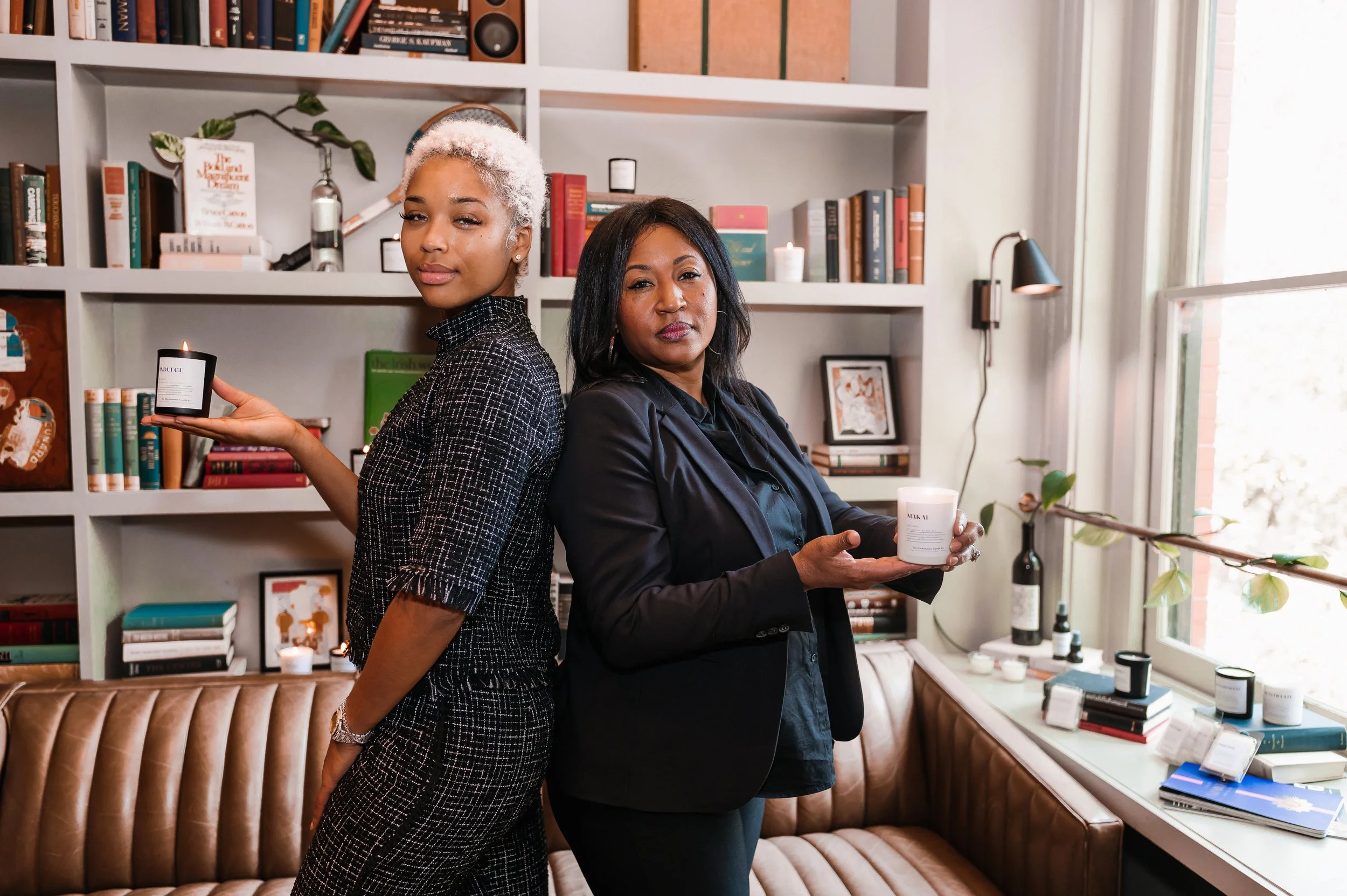 Two women standing back-to-back in a bookstore or library, holding candles and candles in jars, with books and decor on shelves behind them and large windows to the right.