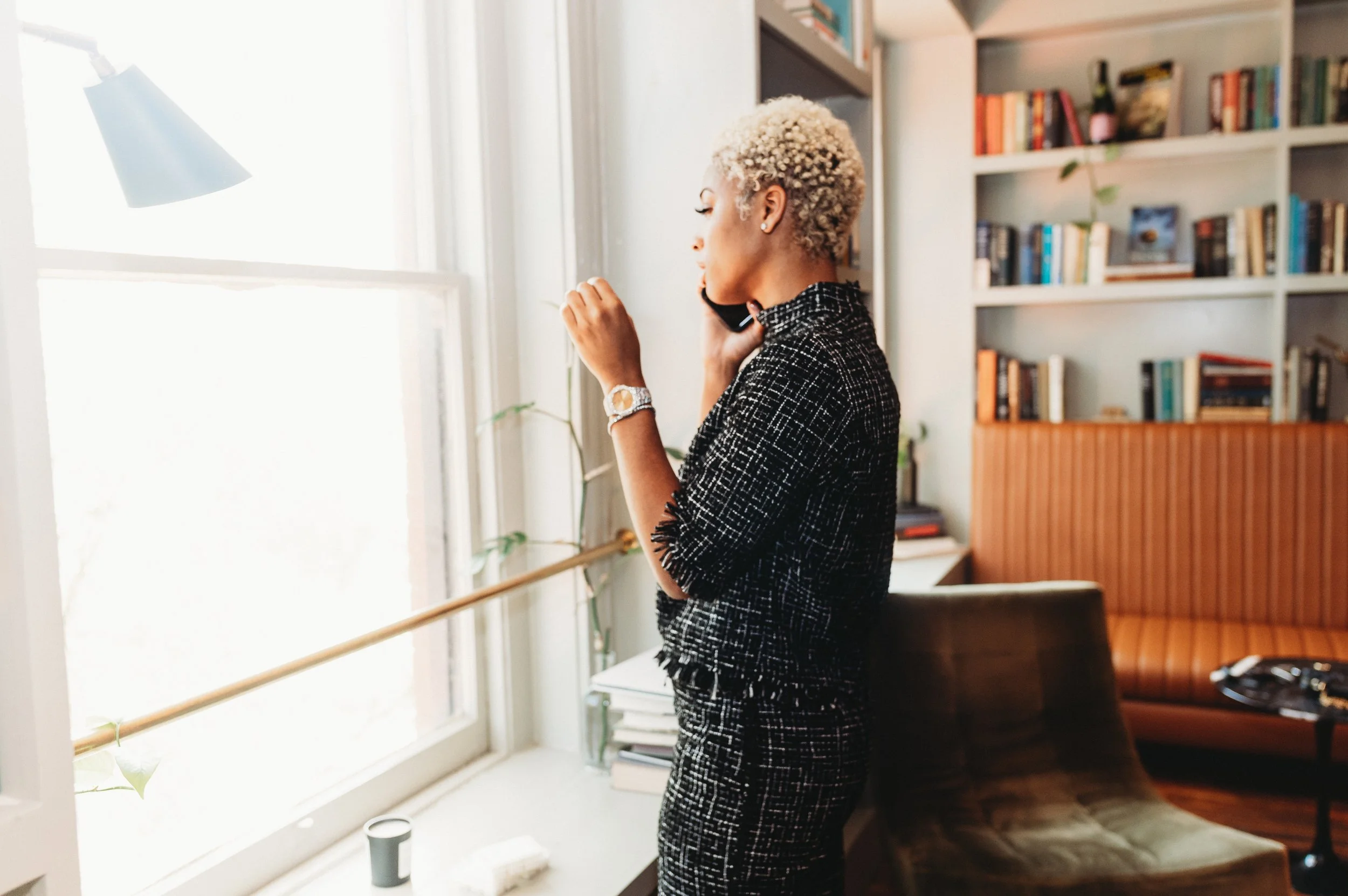 A woman with short, curly, blonde hair is talking on a mobile phone while standing by a large window in a room filled with bookshelves.