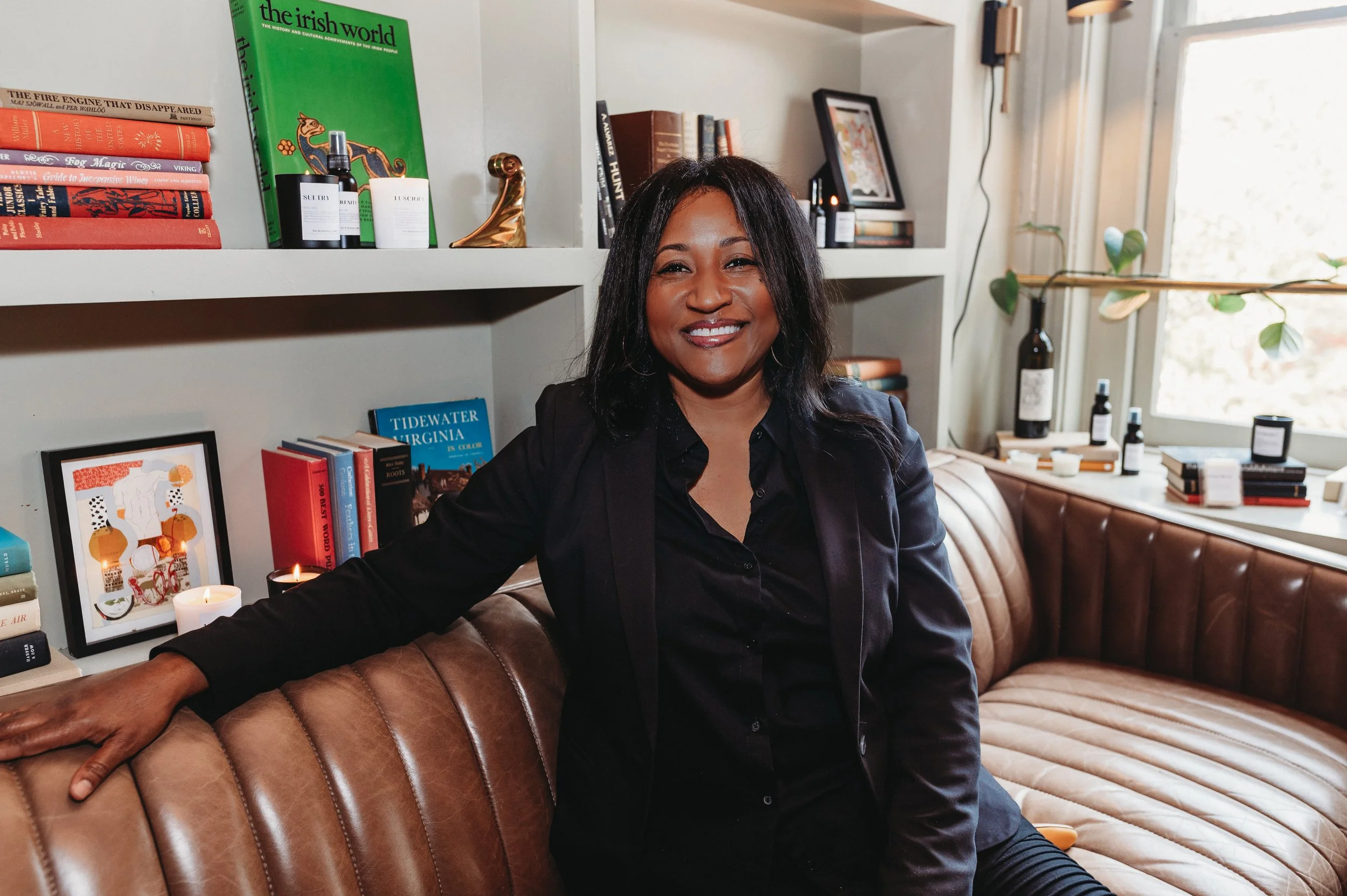 A woman with shoulder-length black hair, smiling, wearing a black blazer and shirt, sitting on a tan leather couch in a well-lit room with books and decorative items on shelves behind her and a window to the right.