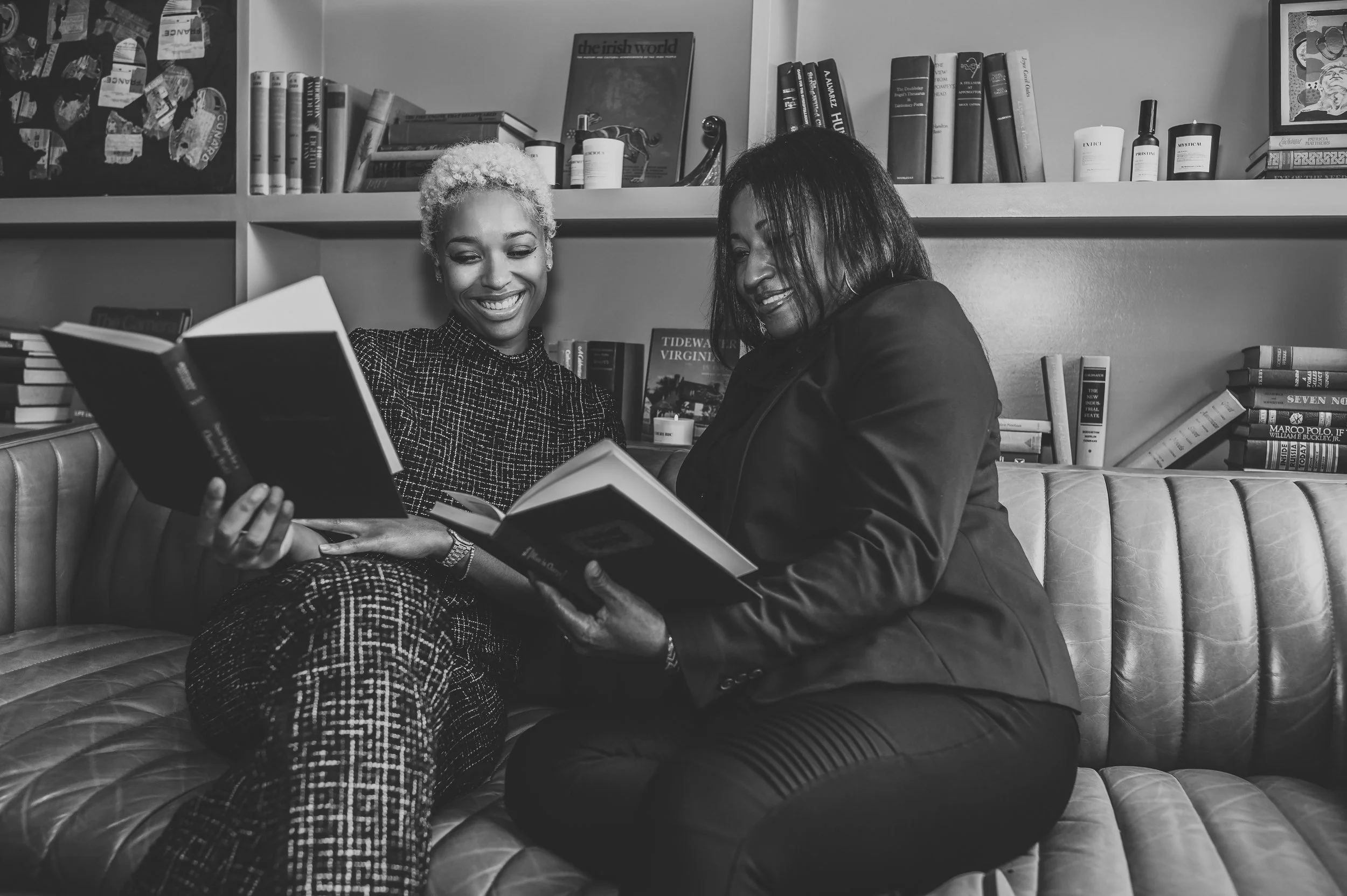 Two women sitting on a leather couch, smiling and reading books together in a room with bookshelves.