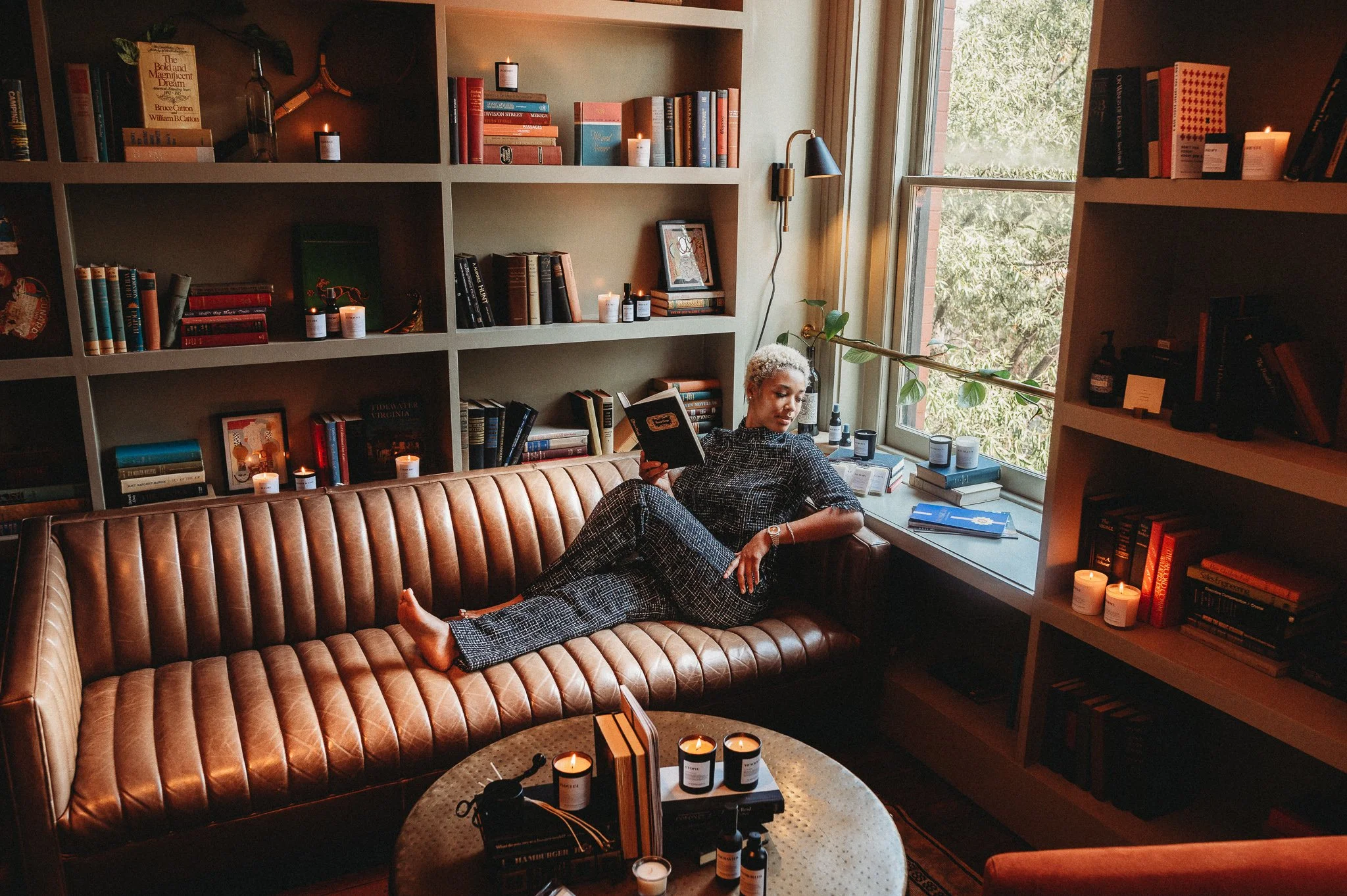 A woman with short curly platinum blonde hair reclining on a brown leather sofa in a cozy room with bookshelves, candles, and a large window with green foliage outside.