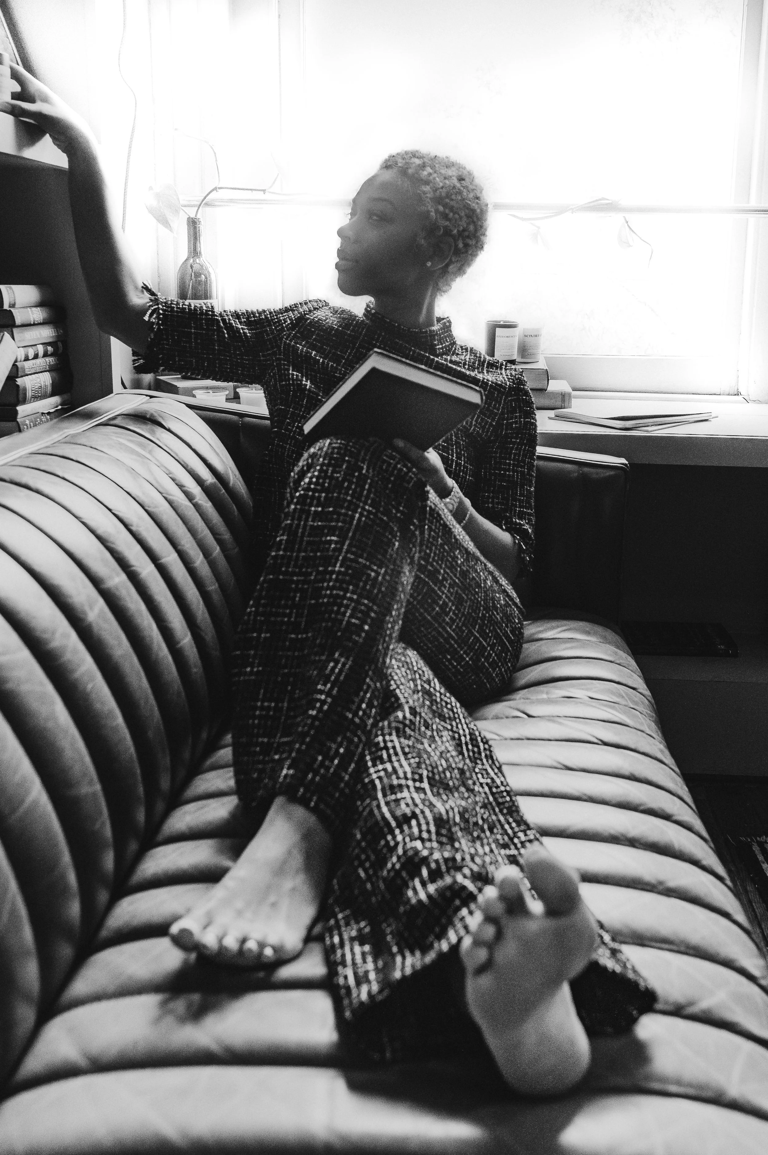 Woman sitting on a leather sofa taking a selfie with a smartphone, holding a book in her other hand, near a window with books and decorative items behind her.