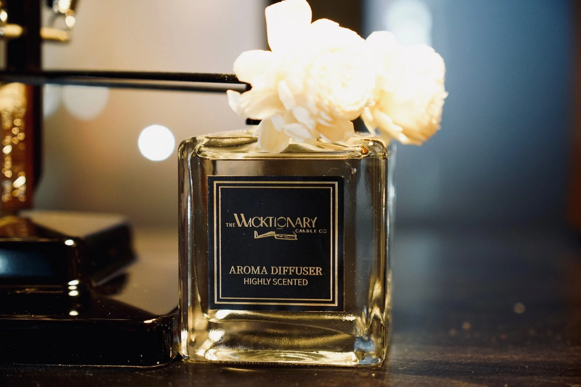 A glass aroma diffuser labeled 'The Wicktory Candle Co' with white flowers on top, placed on a dark surface. Blurred lights in the background.