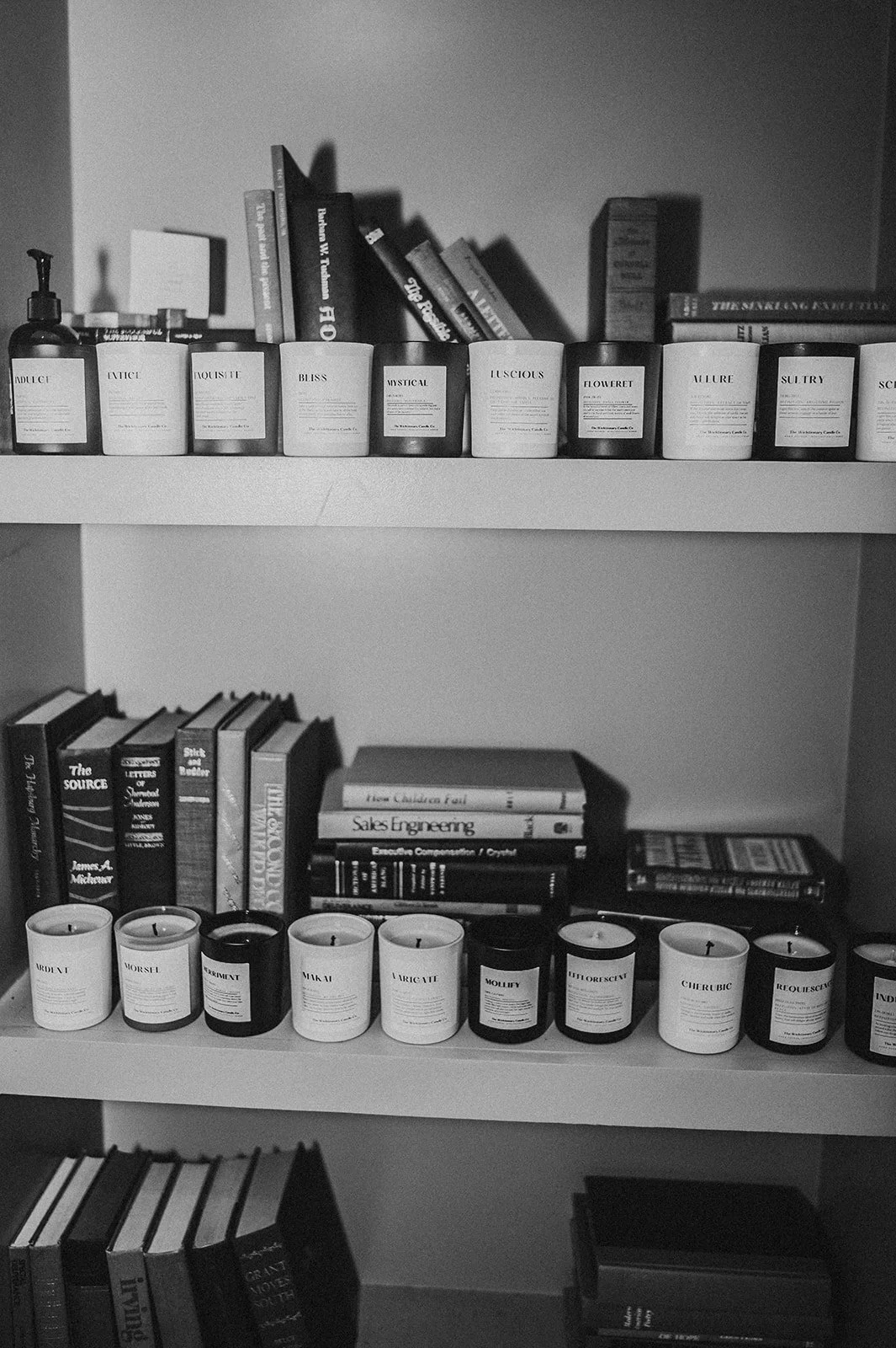 Shelf with candles labeled with various names, books stacked behind, and a small bottle of hand sanitizer on the left.