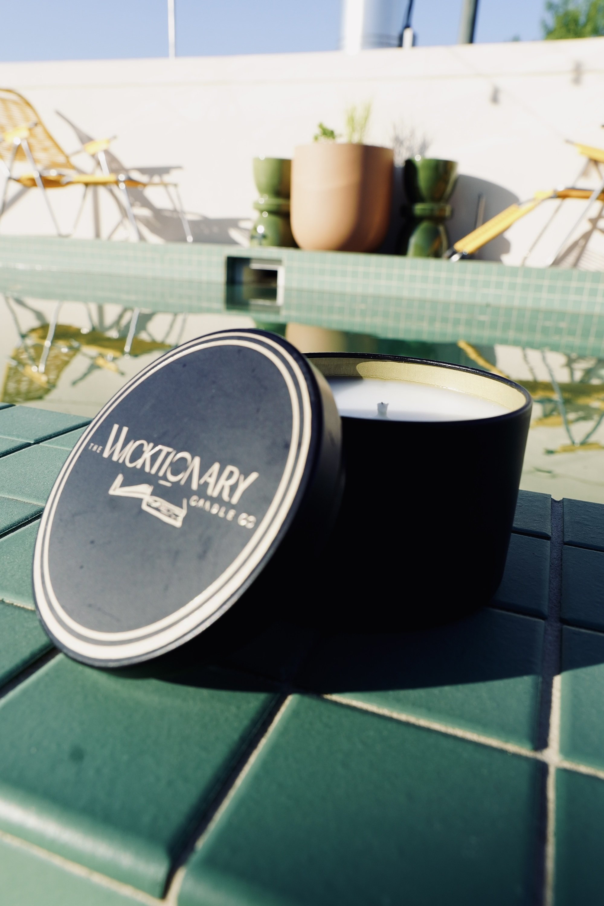 A black candle holder with a white candle inside, placed on a green tiled table outdoors. The candle lid reads 'The Wacktomary, Since 68', and in the background are potted plants and yellow chairs under a bright blue sky.