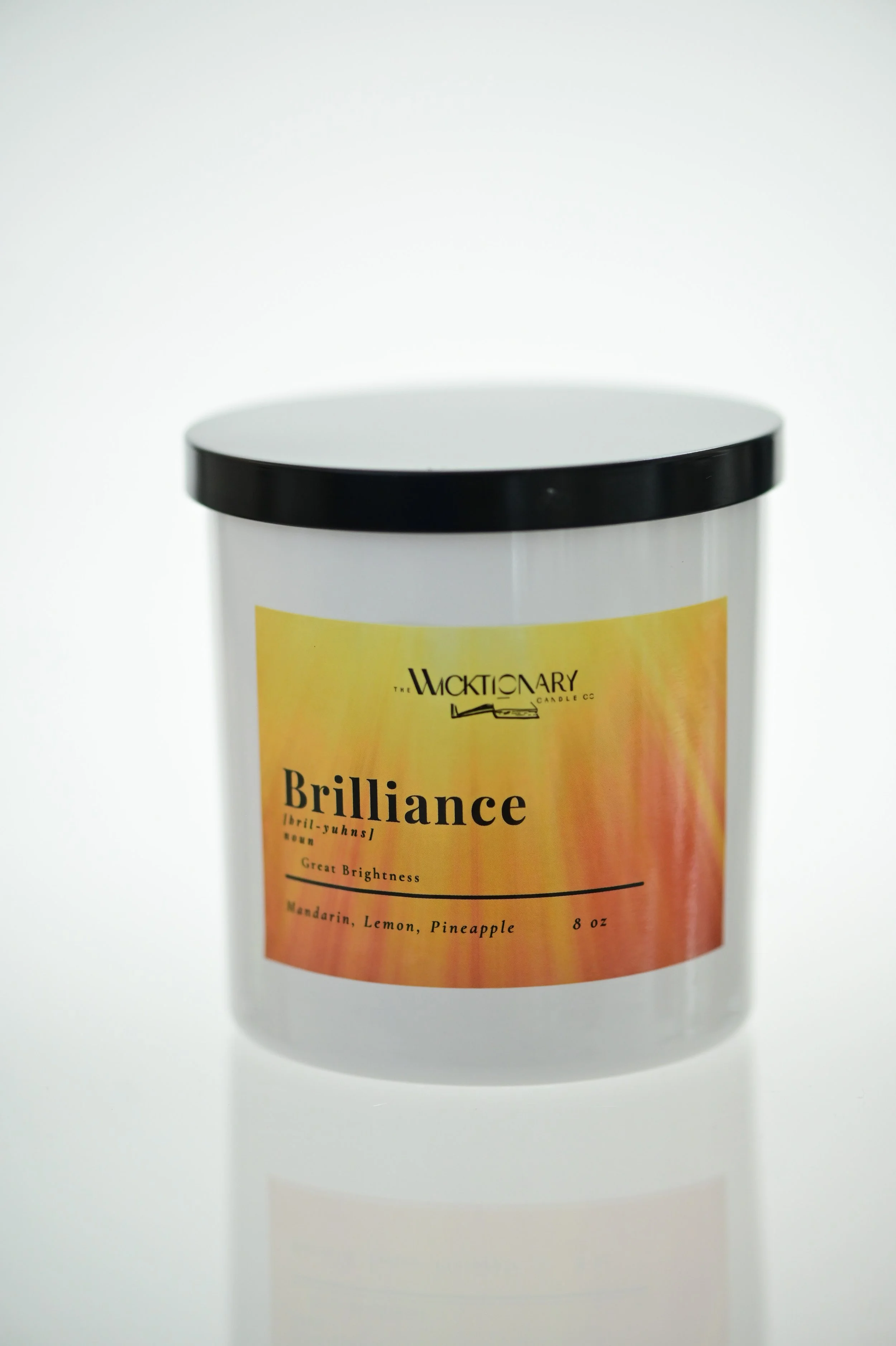 A white candle container with a black lid, labeled 'Brilliance' with a yellow and orange gradient background, and a smaller text mentioning mandarin, lemon, and pineapple scents.