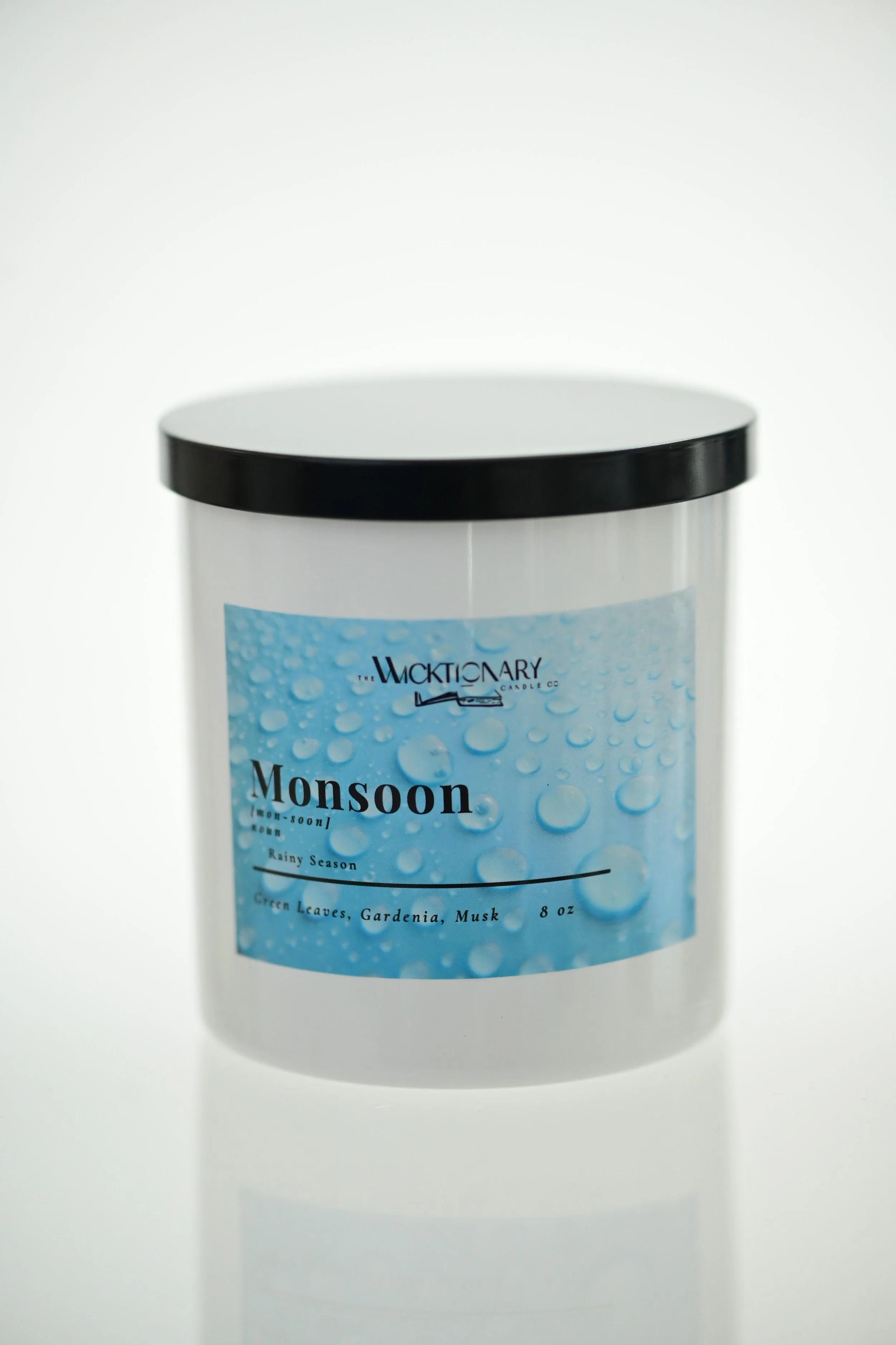 A white candle jar with a black lid, labeled 'Monsoon' with scent notes of green leaves, gardenia, and musk, produced by Wicktionary Candle Co.