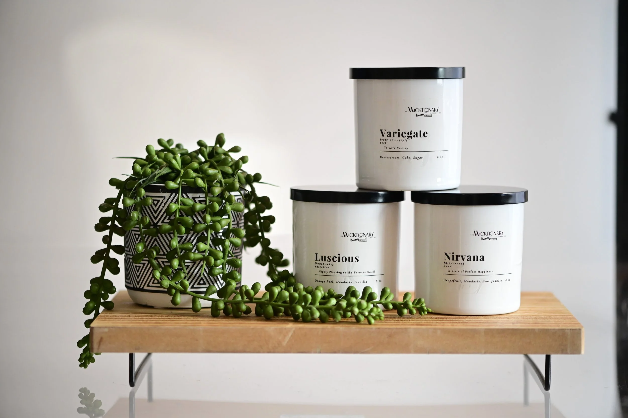 Decorative black and white patterned pot with trailing green succulent, three white jars labeled 'Variegate,' 'Luscious,' and 'Nirvana' with black lids on a wooden shelf.