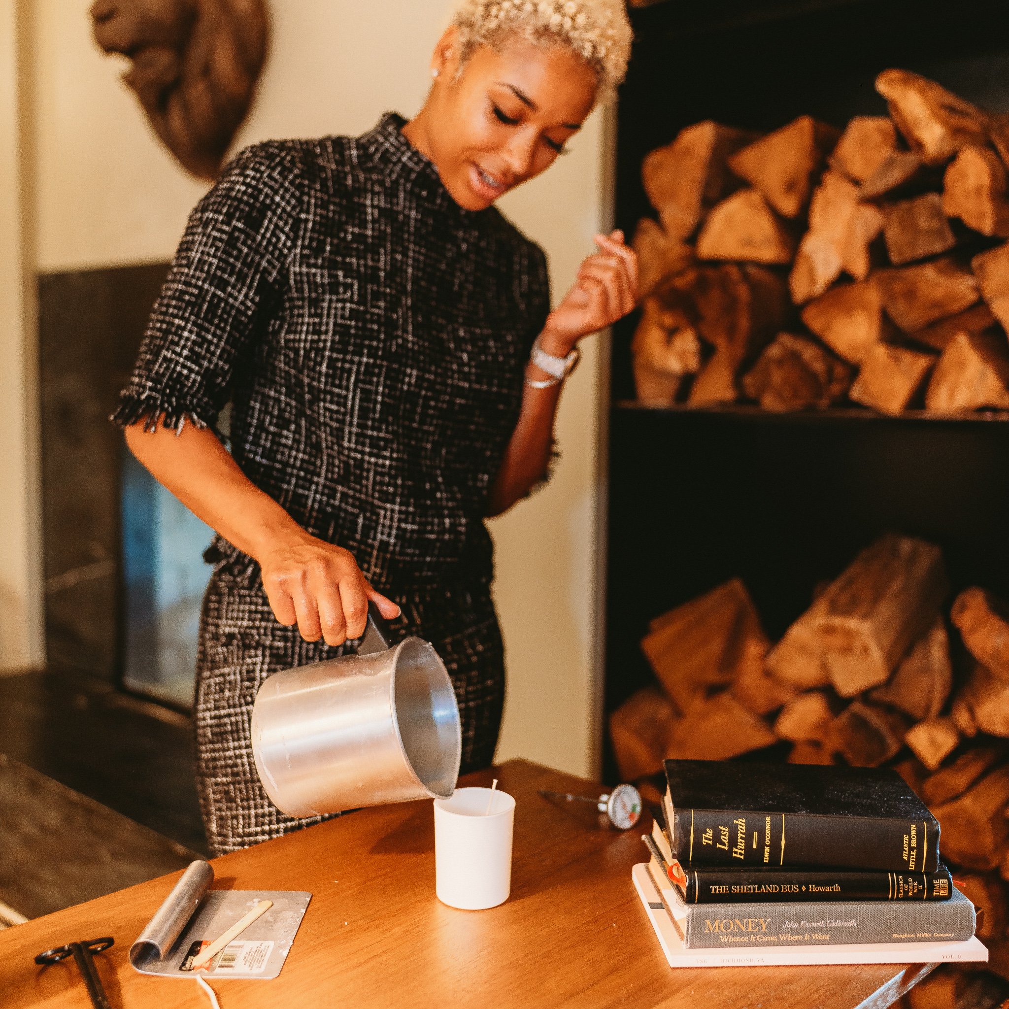 Woman with short blonde curly hair and checkered dress pouring wax into a candle mold on a wooden table with books, a thermometer, and a metal tool nearby, in front of a stacked firewood display.