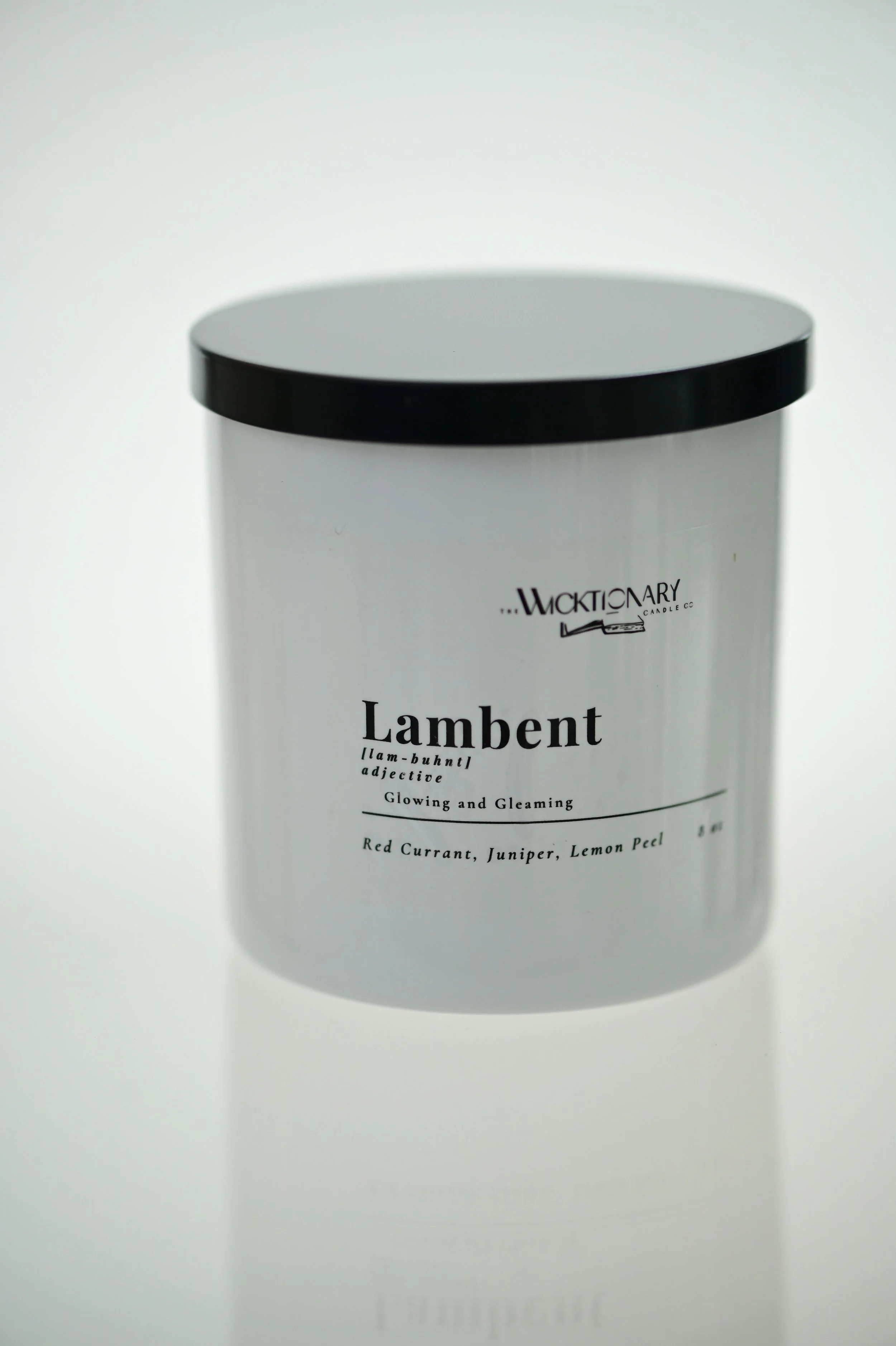 A white container with a black lid, labeled 'Lambent', a scented candle with notes of red currant, juniper, and lemon peel.