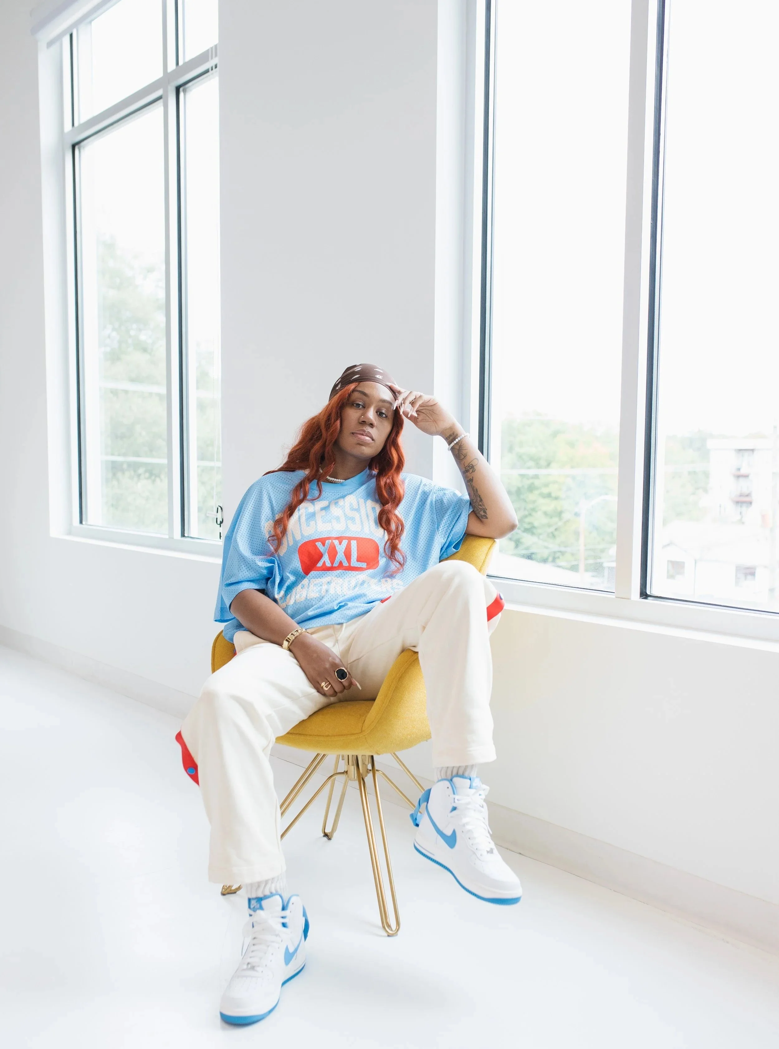 A woman with red hair sitting on a yellow chair next to large windows in a bright room. She is wearing a blue oversized shirt, off-white pants, and white sneakers with blue accents, and looking at the camera.
