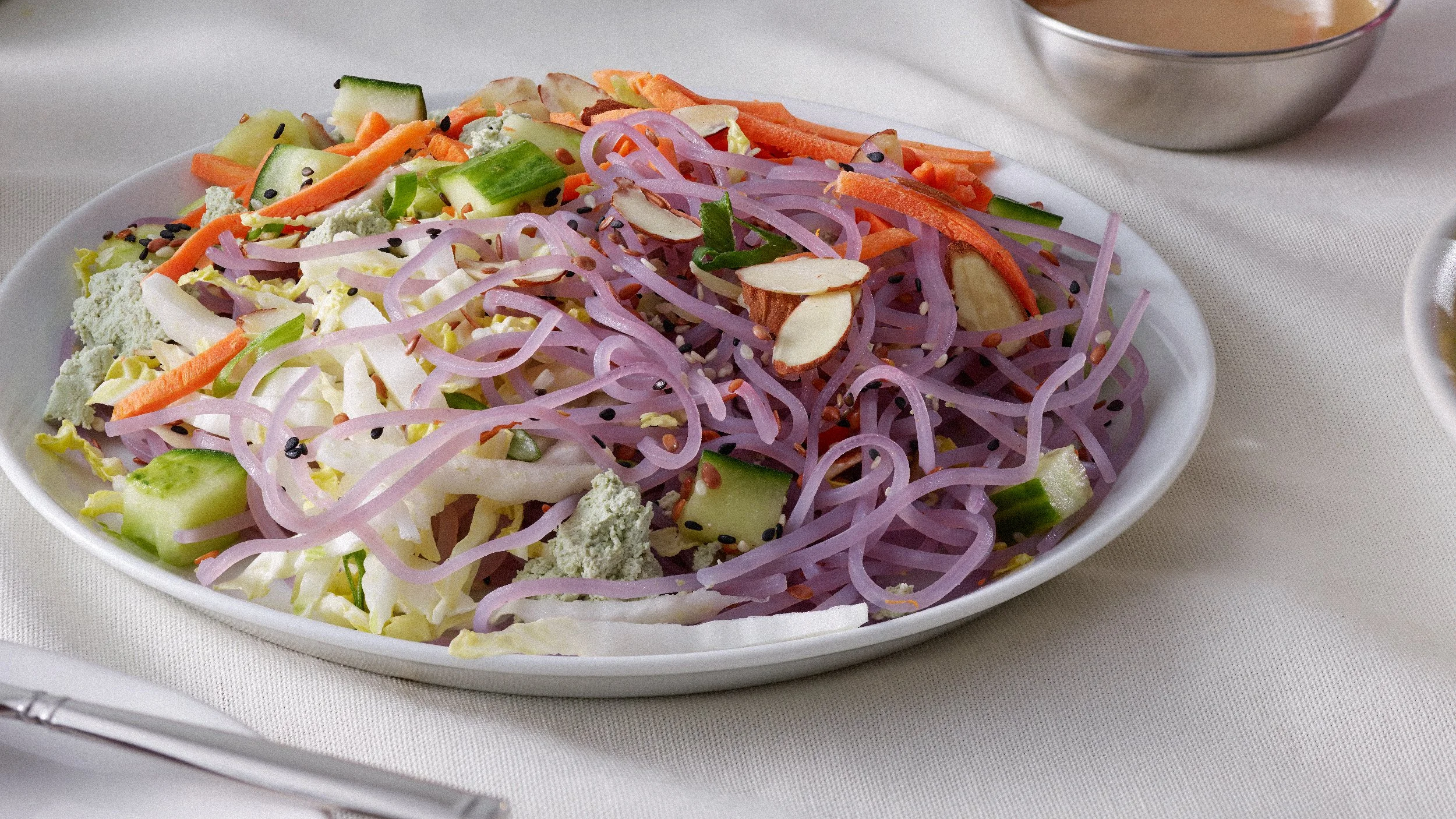A white oval dish filled with a colorful vegetable salad containing shredded purple cabbage, shredded carrots, chopped cucumber, sliced almonds, crumbled cheese, and black sesame seeds on a white linen tablecloth.