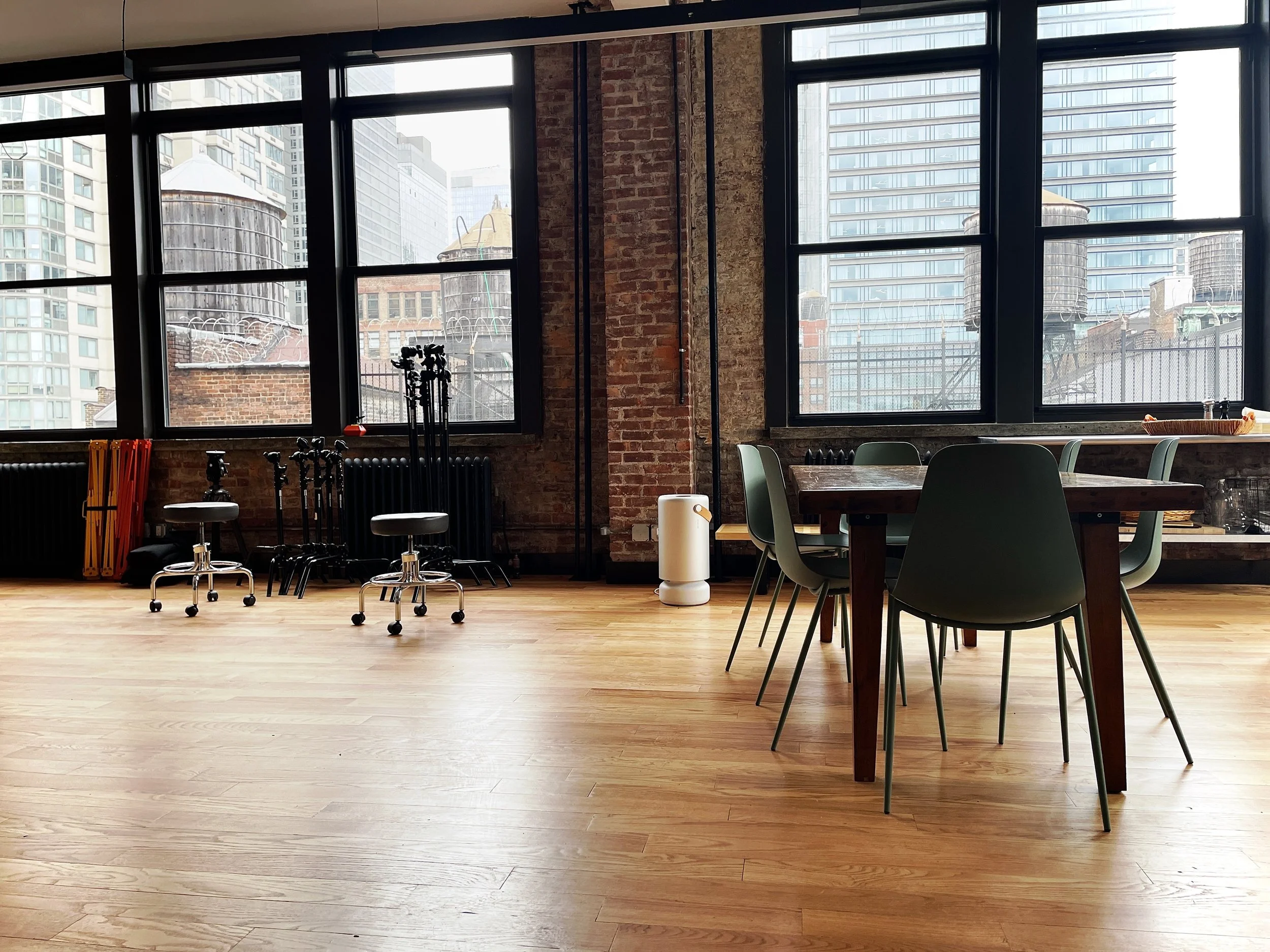 Interior of a modern, industrial-style room with large windows, a wooden floor, a table with six chairs, and some equipment stored near the windows.