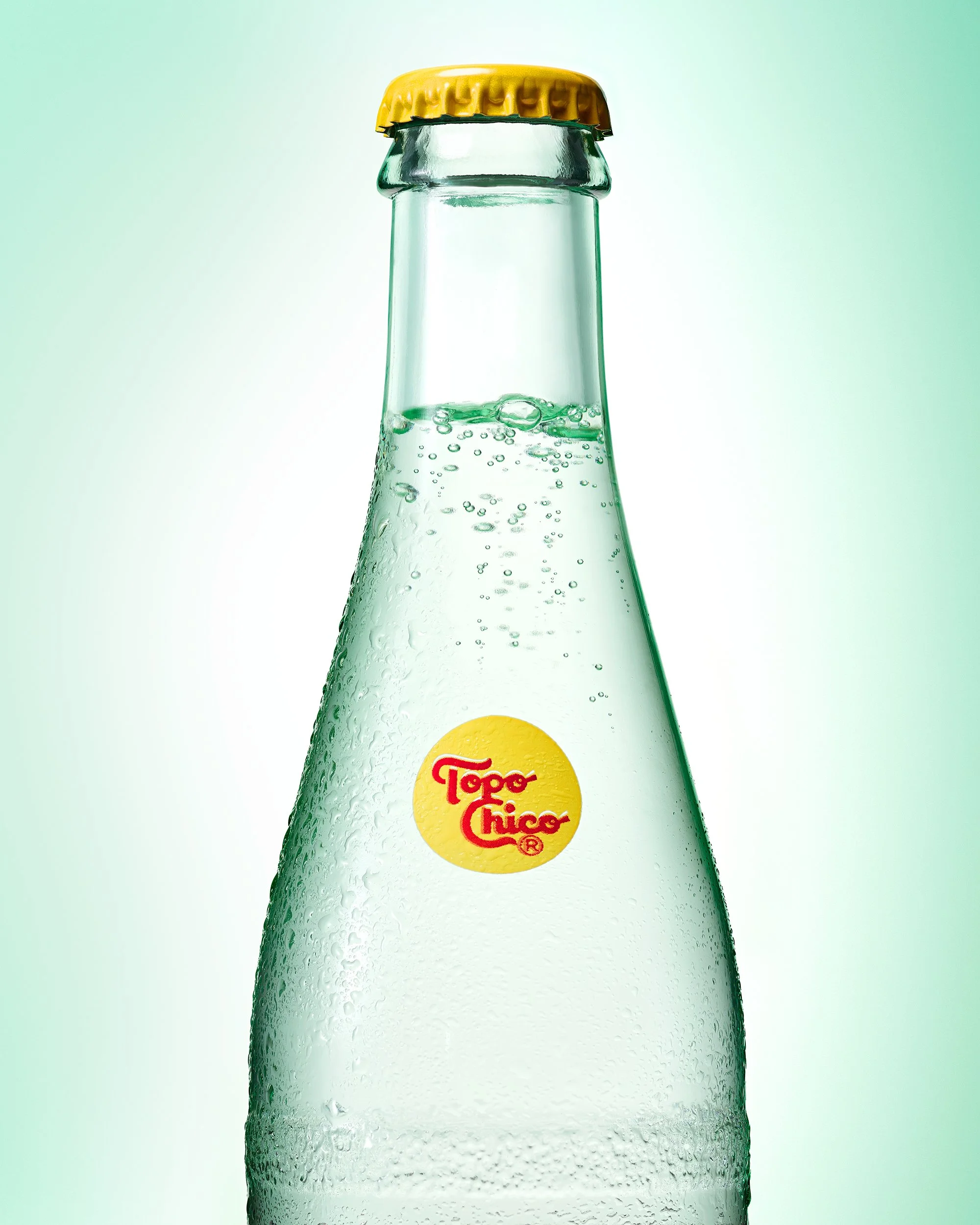 A single glass bottle of Topo Chico mineral water is shown with condensation droplets against a monchromatic light green halo background