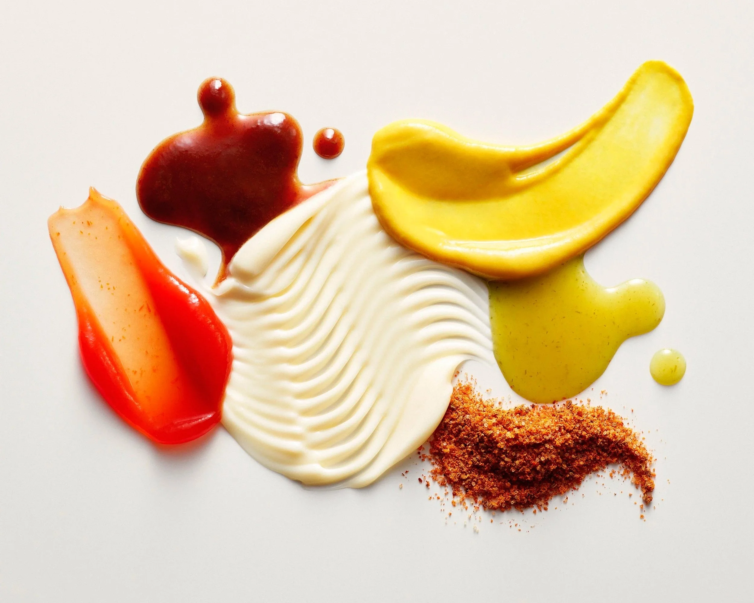 Ketchup, mustard, mayonnaise, hot sauce and spice rub are arranged and stylized similar to cosmetic textures on a warm white background