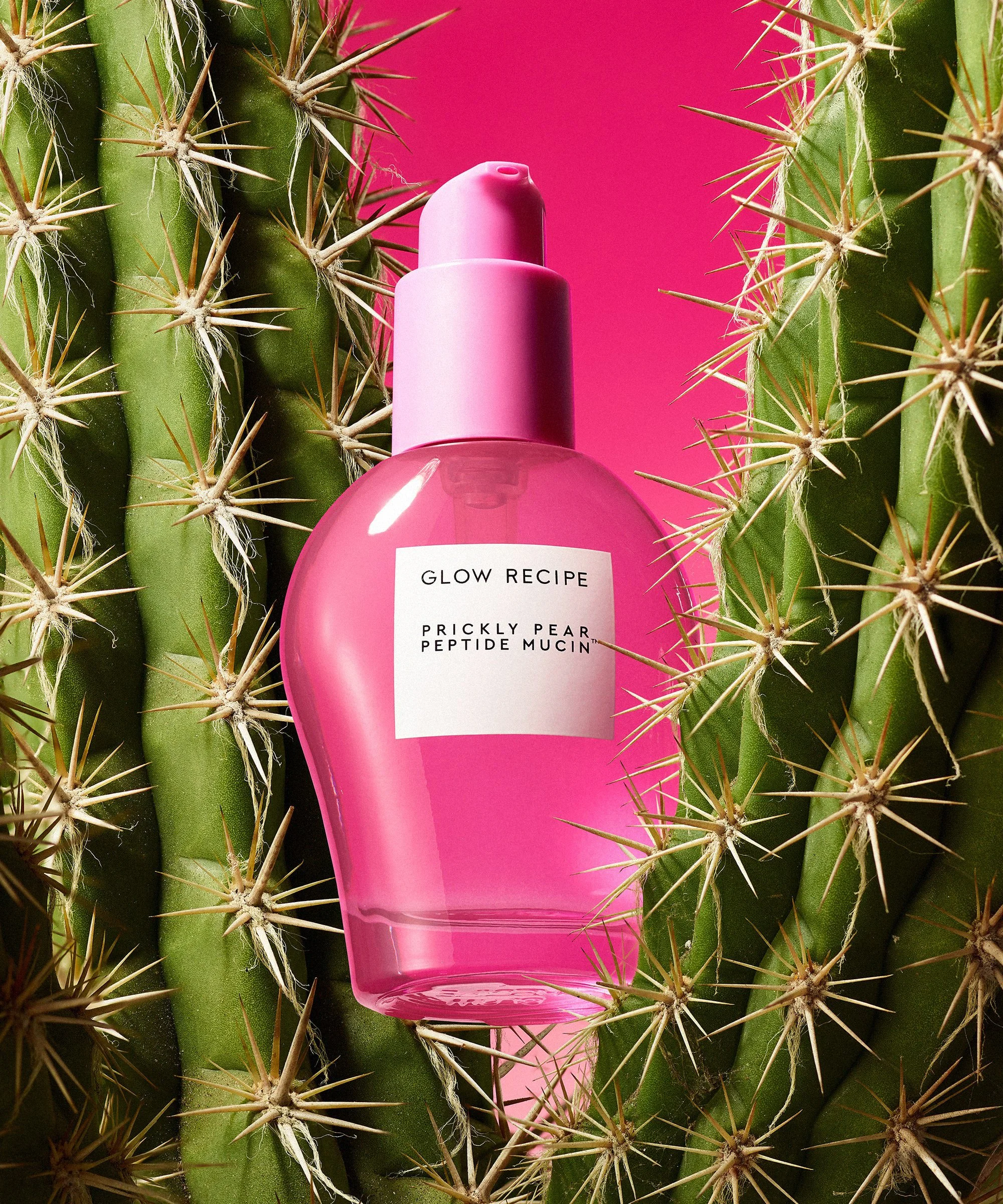 A bright pink glass serum skincare bottle sits wedged between two prickly cactus branches. The background shows a deep pink gradient