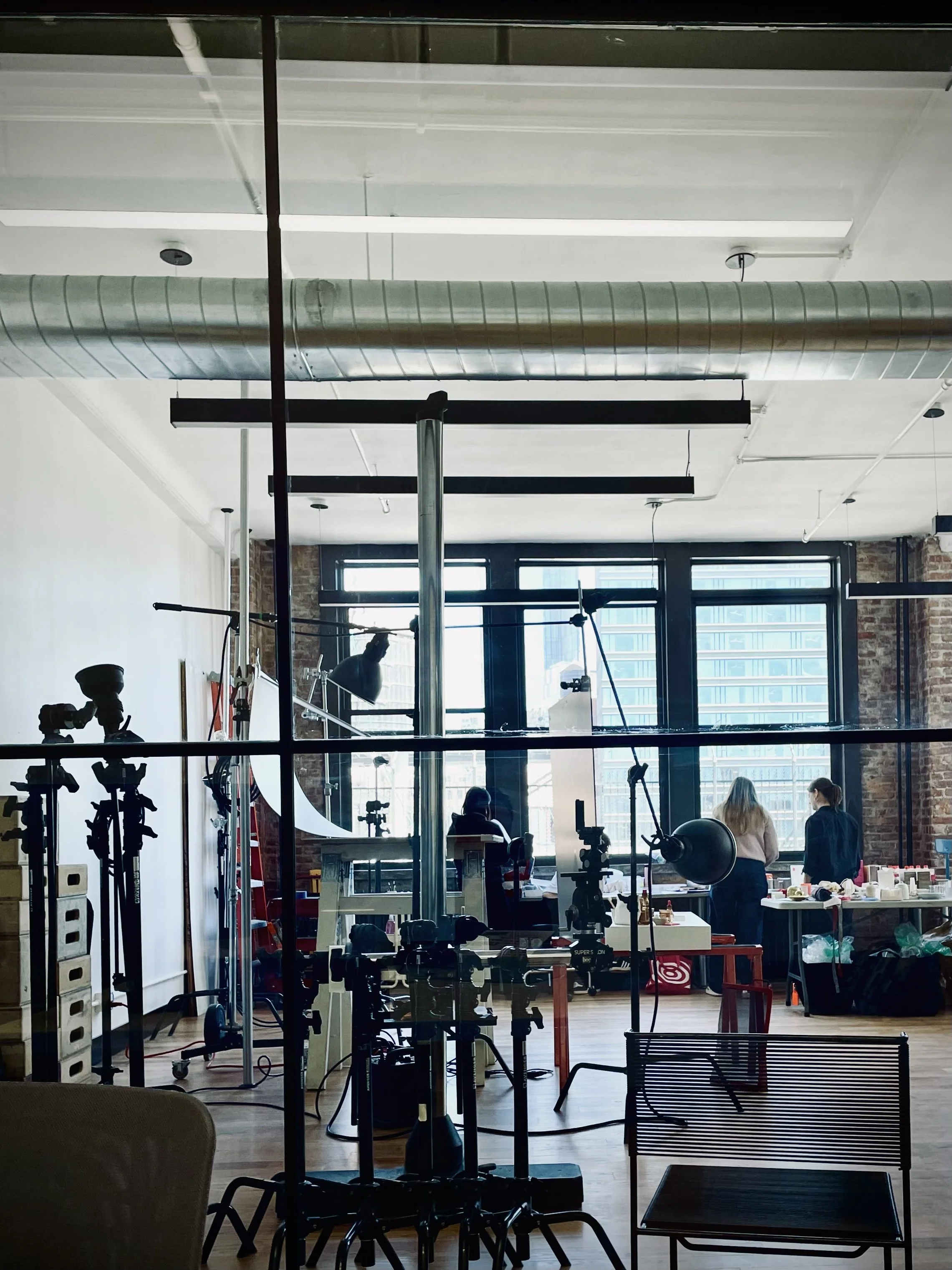 Photo of a photography studio with cameras, tripods, and lighting equipment set up near large windows, with three people working at tables in the background.