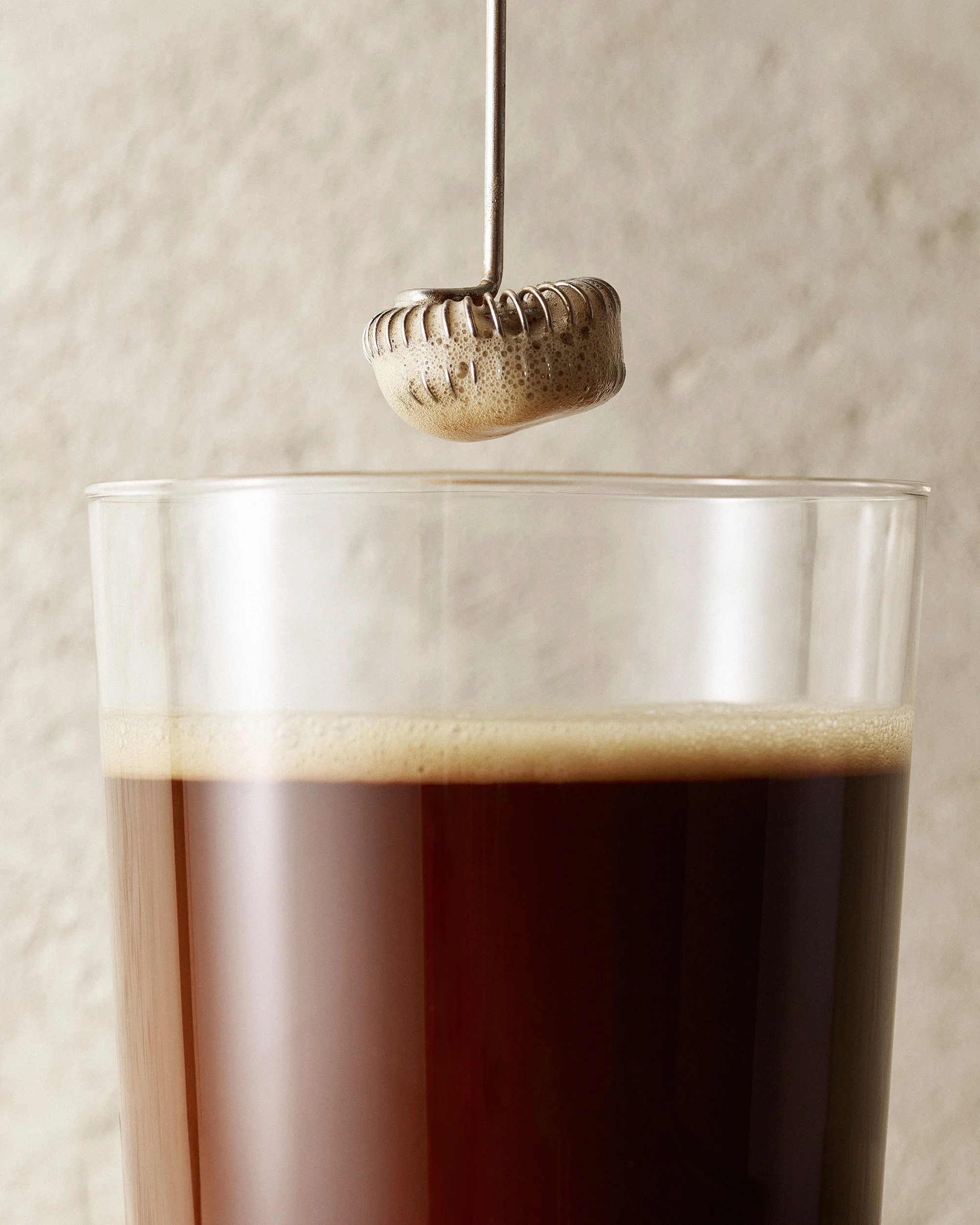 A metal frother tool containing foam hovers above a clear drinking glass with coffee against a textured stone background