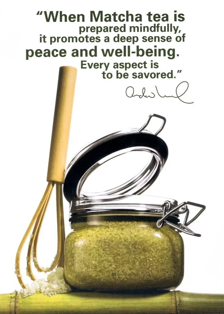 A glass jar of green skin scrub is shown sitting on a shelf of green bamboo next to a wooden whisk against a white background